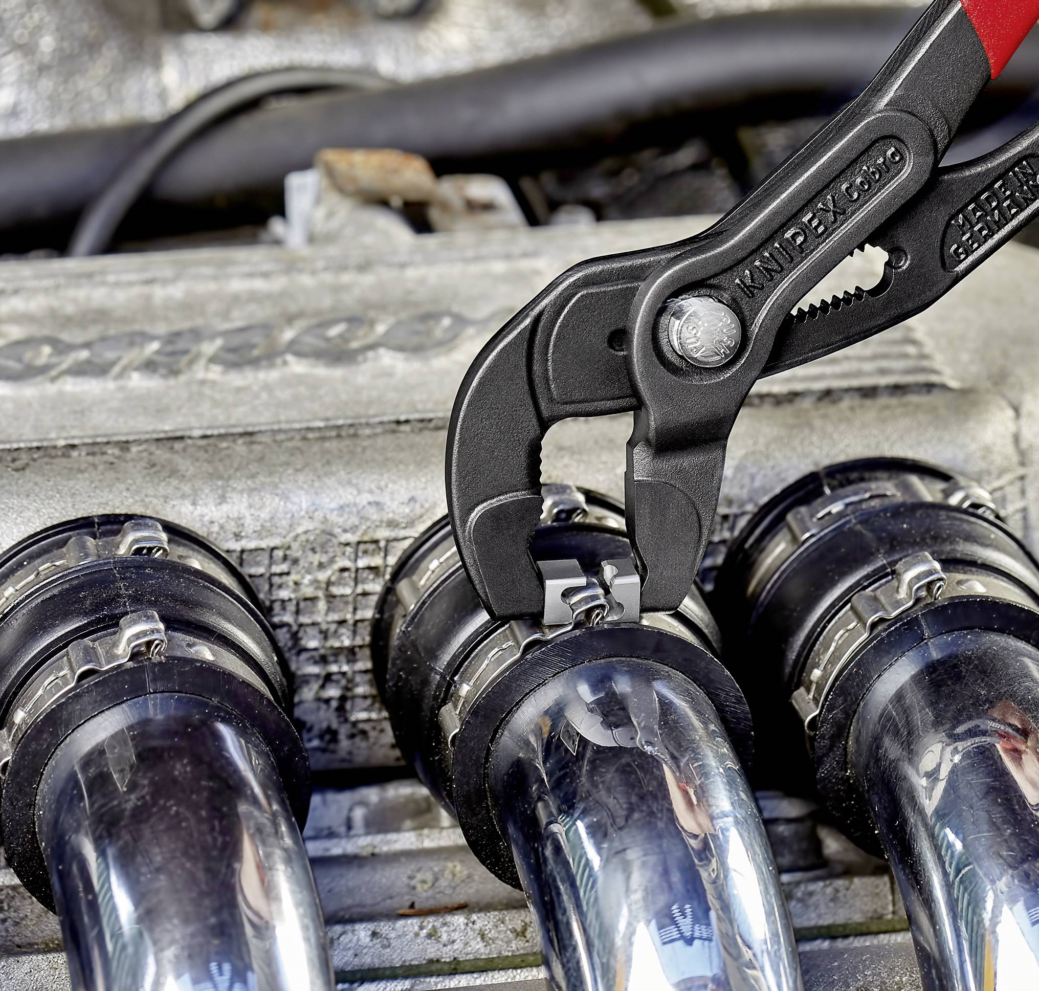 Eine Knipex-Zange löst eine Schlauchklemme an einem Autokühlersystem. Nahe Ansicht mit Fokus auf das Werkzeug und die Schlauchverbindungen.