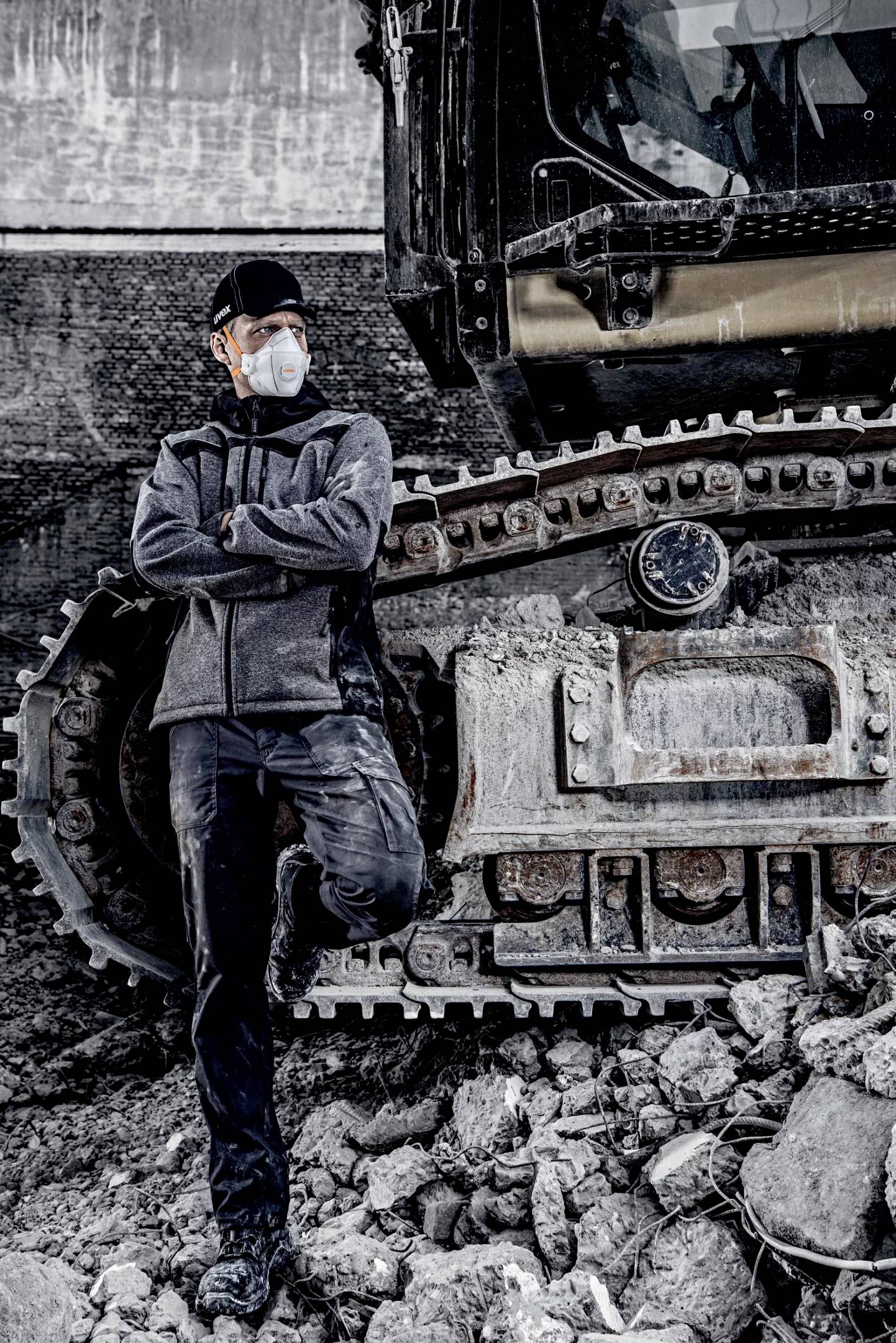 Ein Arbeiter mit Gesichtsmaske lehnt an einem großen Bagger in einer Baustelle. Er trägt dunkle Arbeitskleidung und ist staubbedeckt.
