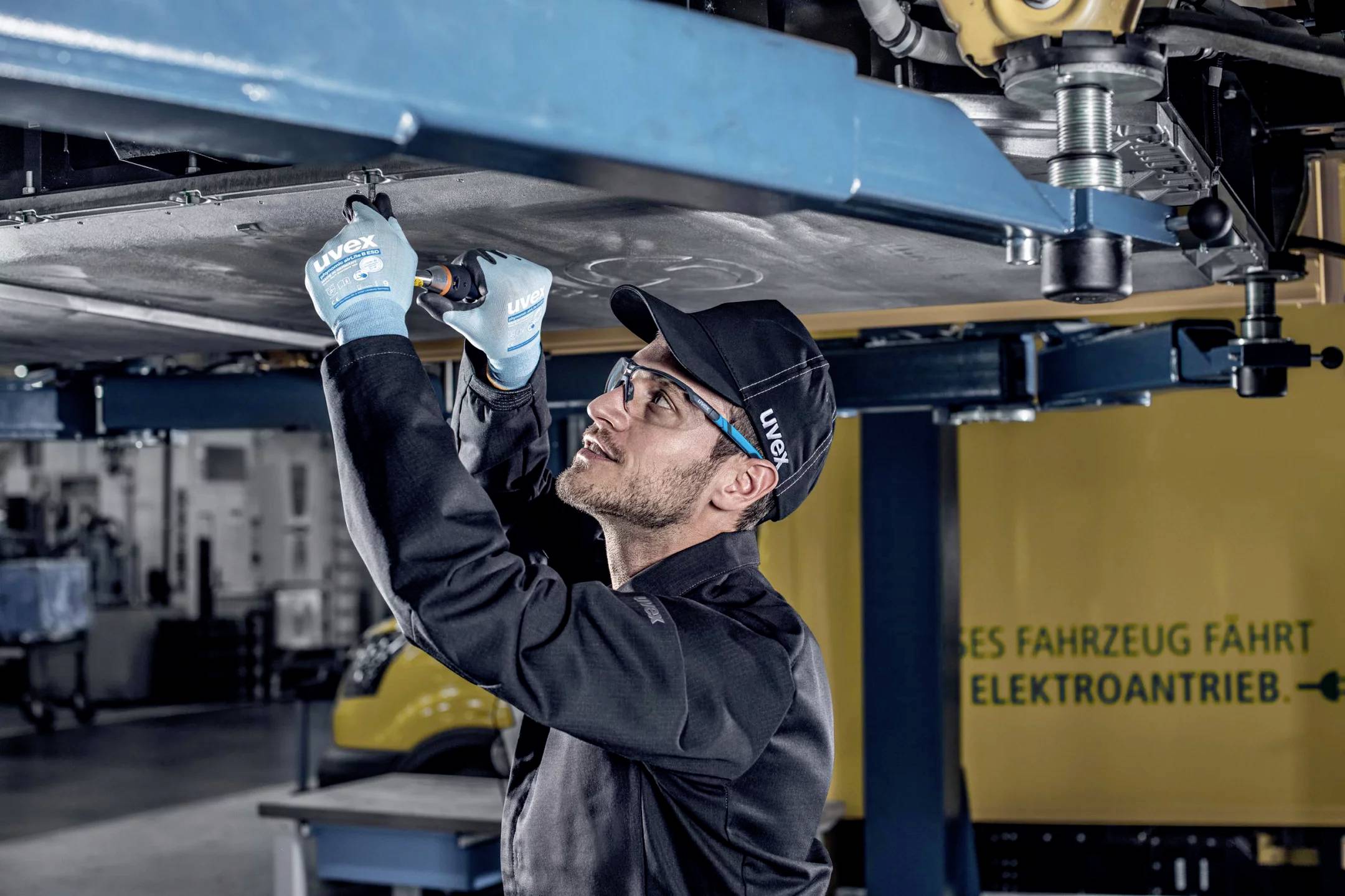 Eine Person in Arbeitskleidung arbeitet unter einem Fahrzeug auf einer Hebebühne in einer Werkstatt.