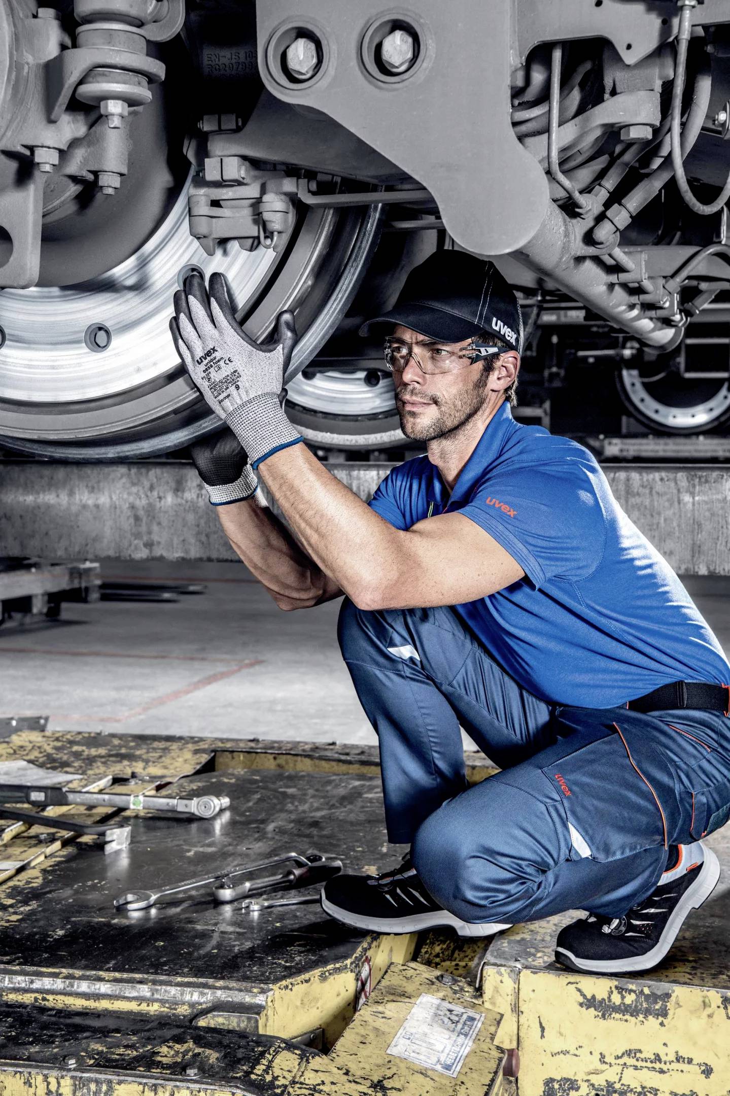 Ein Mechaniker in blauer Arbeitskleidung überprüft das Fahrwerk eines Zuges. Er trägt Handschuhe und sorgfältig schaut auf die Mechanik.