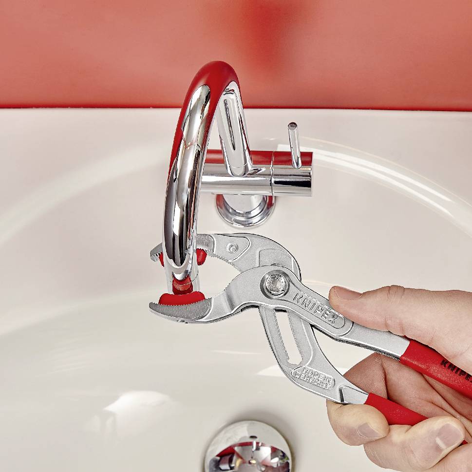 Eine Person repariert mit einer roten Zange einen Wasserhahn in einem Bad. Der Wasserhahn ist in einem Becken montiert.
