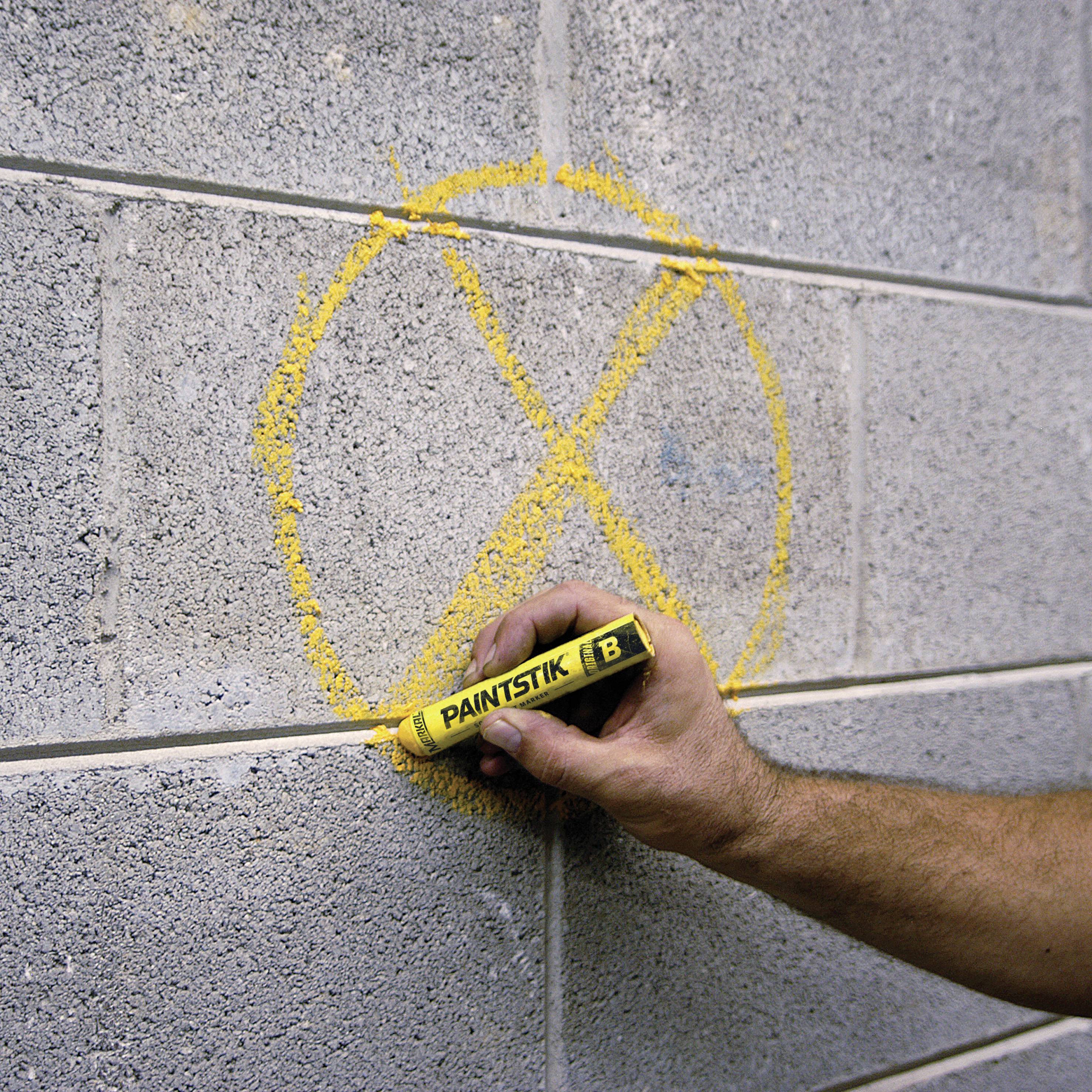 Hand zeichnet mit gelbem Paintstik ein großes X auf eine graue Ziegelwand.