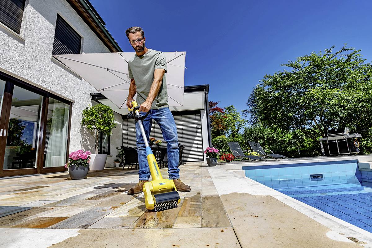 Mann reinigt Terrasse mit gelbem Hochdruckreiniger neben Pool an sonnigem Tag.