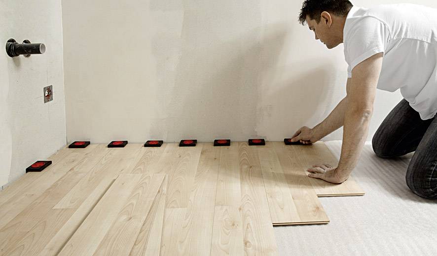 Eine Person verlegt Holzbodenplatten in einem Raum, verwendet Abstandshalter an der Wand zur genauen Ausrichtung.