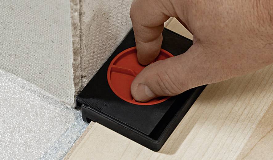 Hand drückt roten Abstandshalter zwischen Wand und Holzfußboden zur Sicherstellung eines gleichmäßigen Spalts bei der Installation.
