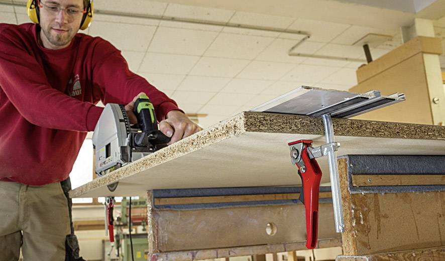 Ein Mann arbeitet mit einer Elektrosäge, um eine Holzplatte auf einem Tisch zuzuschneiden. Er trägt Schutzbrille und Gehörschutz.