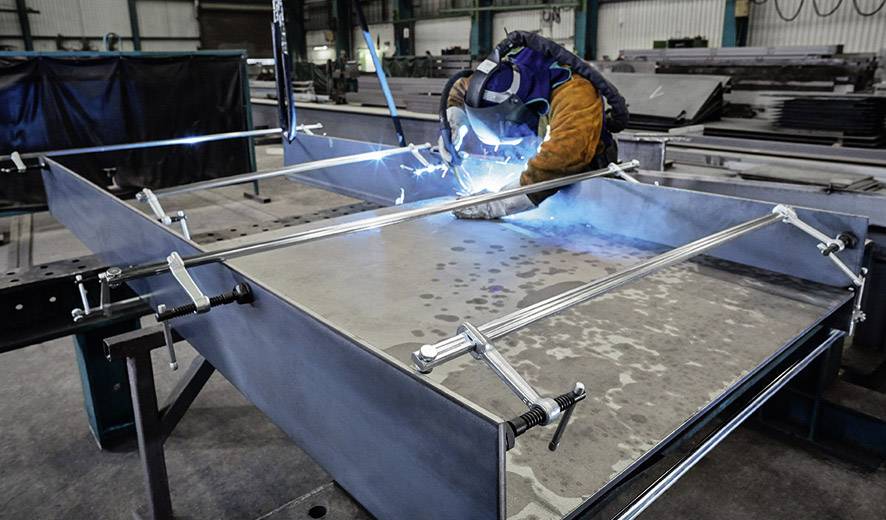 Ein Schweißer in Schutzkleidung arbeitet in einer Werkstatt an einem Metallstück, umgeben von Klammern und Geräten, Lichtblitze sichtbar.
