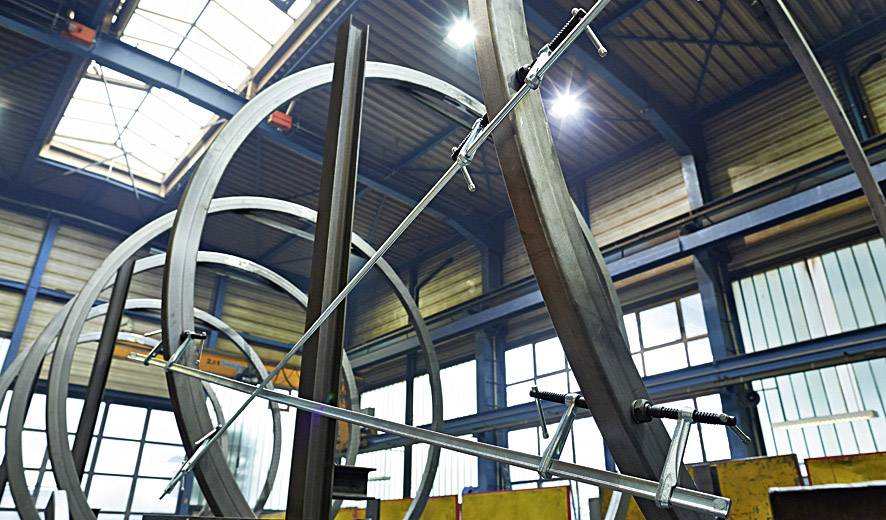 Stählerne Skulptur im Bau in einer Werkhalle mit sichtbarer Dachkonstruktion und Lichteinfall; Metallbalken in runden Formen.