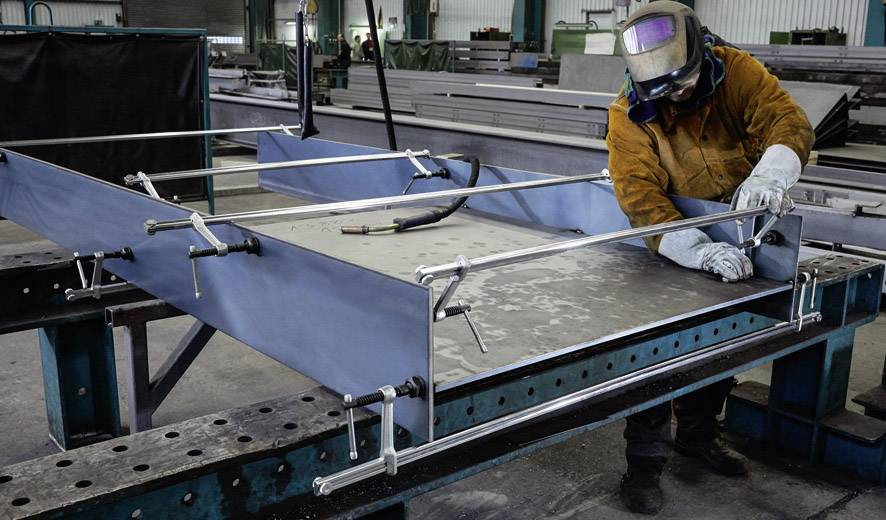 Eine Person mit Schutzkleidung und Schweißhelm arbeitet an einer Metallkonstruktion in einer Fabrikhalle.