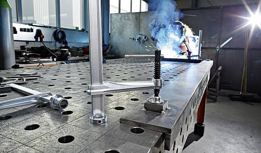 Eine Person schweißt an einem Metalltisch in einer Werkstatt. Funken sprühen, und der Raum ist mit Werkzeugen ausgestattet.