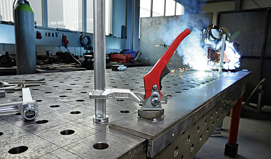 Metallbearbeitungswerkstatt mit einem Schweißer im Hintergrund, der funkenreizend arbeitet. Eine Metallklemme im Vordergrund.