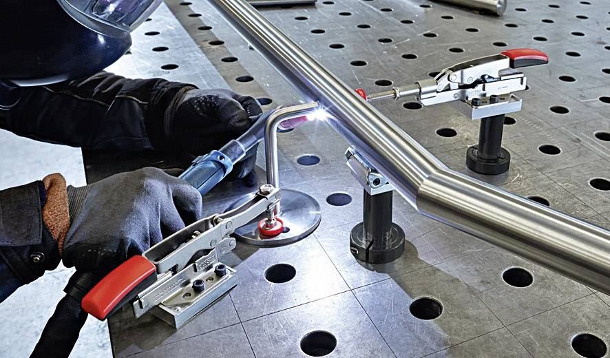 Eine Person führt Schweißarbeiten an einem Metallrohr auf einem Metalltisch durch, gesichert mit Klemmen.