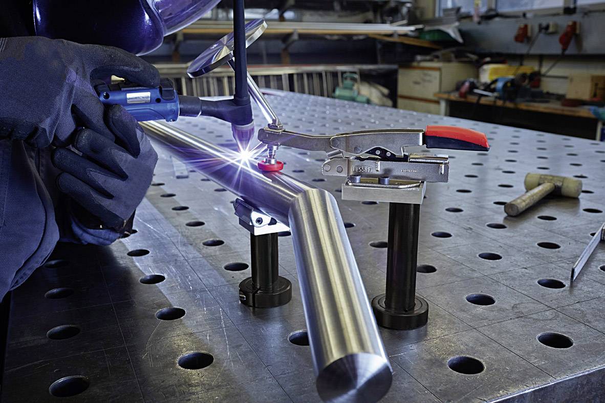 Eine Person schweißt Metallrohr auf einem Werkstatt-Arbeitstisch mit Schutzkleidung und Werkzeugen im Hintergrund.