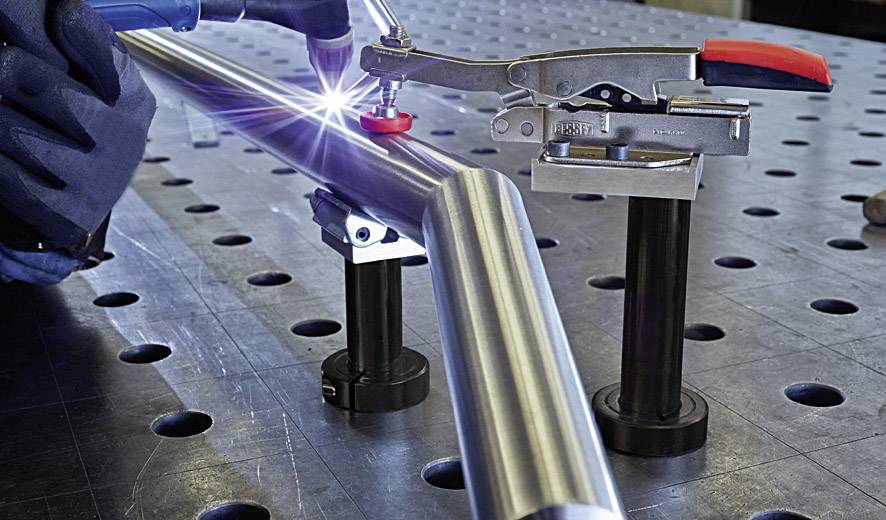 Nahaufnahme eines Schweißvorgangs: Ein Arbeiter schweißt ein Metallrohr auf einem Werkstatttisch.