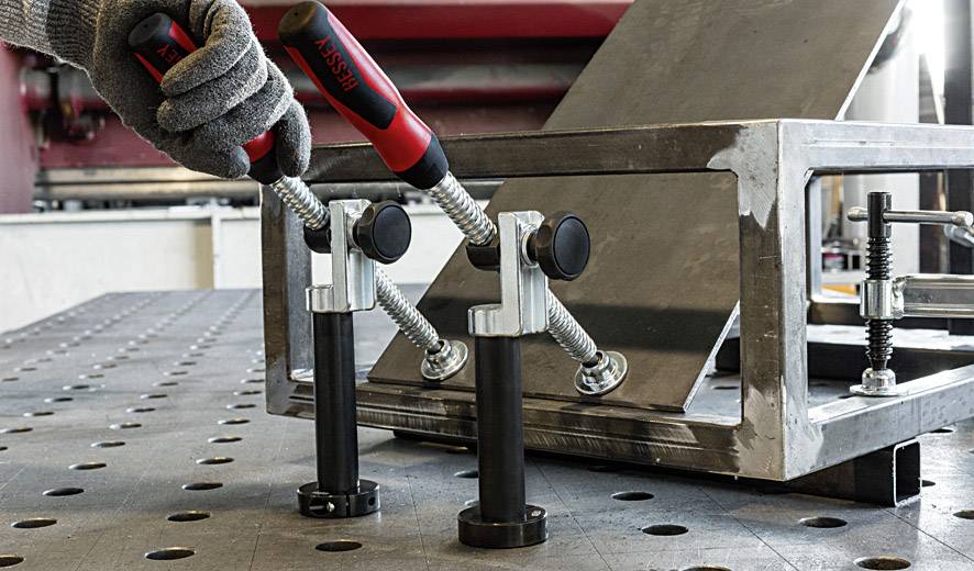 Eine Hand in einem Arbeitshandschuh justiert Metallklemmen auf einem Werkstatttisch, um ein Metallstück in Position zu halten.