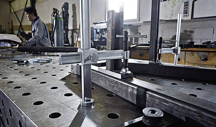 Ein Metallarbeiter benutzt einen Schweißtisch mit Spannwerkzeugen in einer Werkstatt. Im Hintergrund stehen weitere Werkzeugmaschinen.