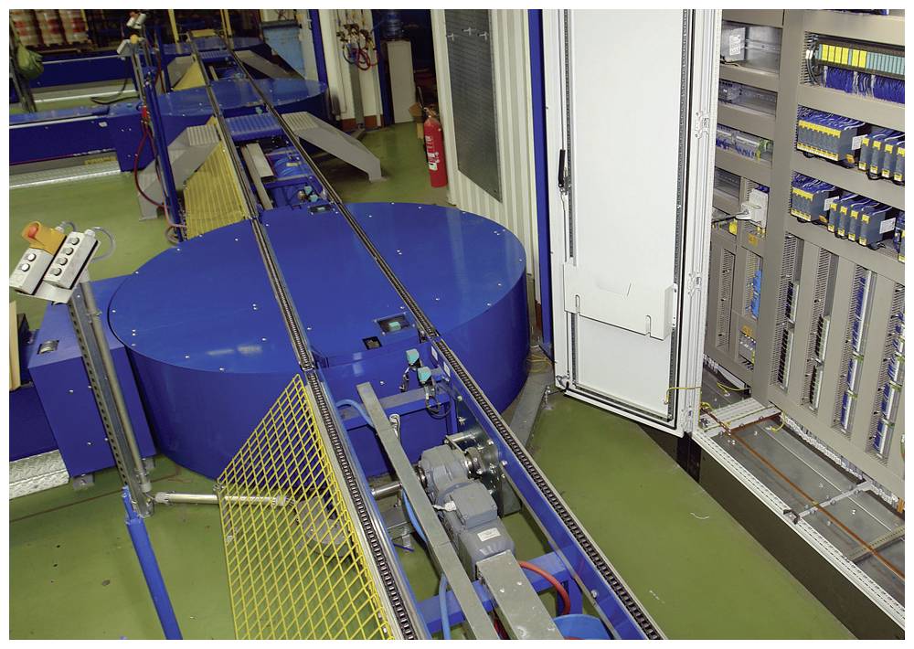 Produktionsanlage mit blauer Drehscheibe und Förderband, umgeben von Schaltkästen und Sicherheitsausrüstung.