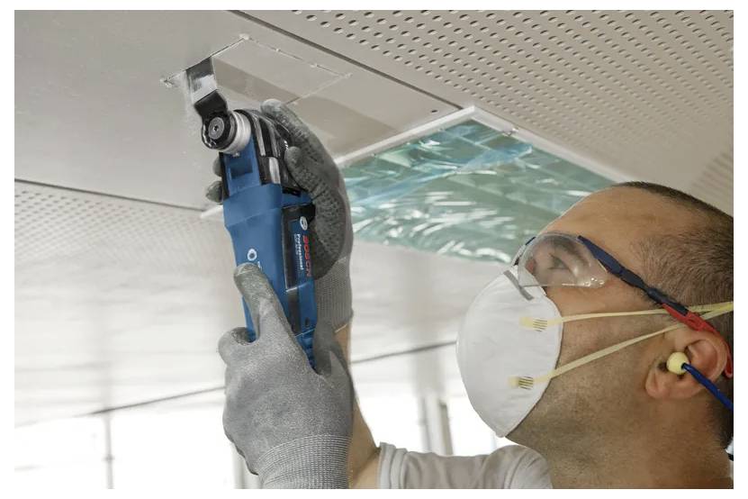 Techniker mit Handschuhen, Maske und Schutzbrille inspiziert einen Deckenbelüftungsschacht oder ein Deckenpaneel mit einem blauen tragbaren Gerät.