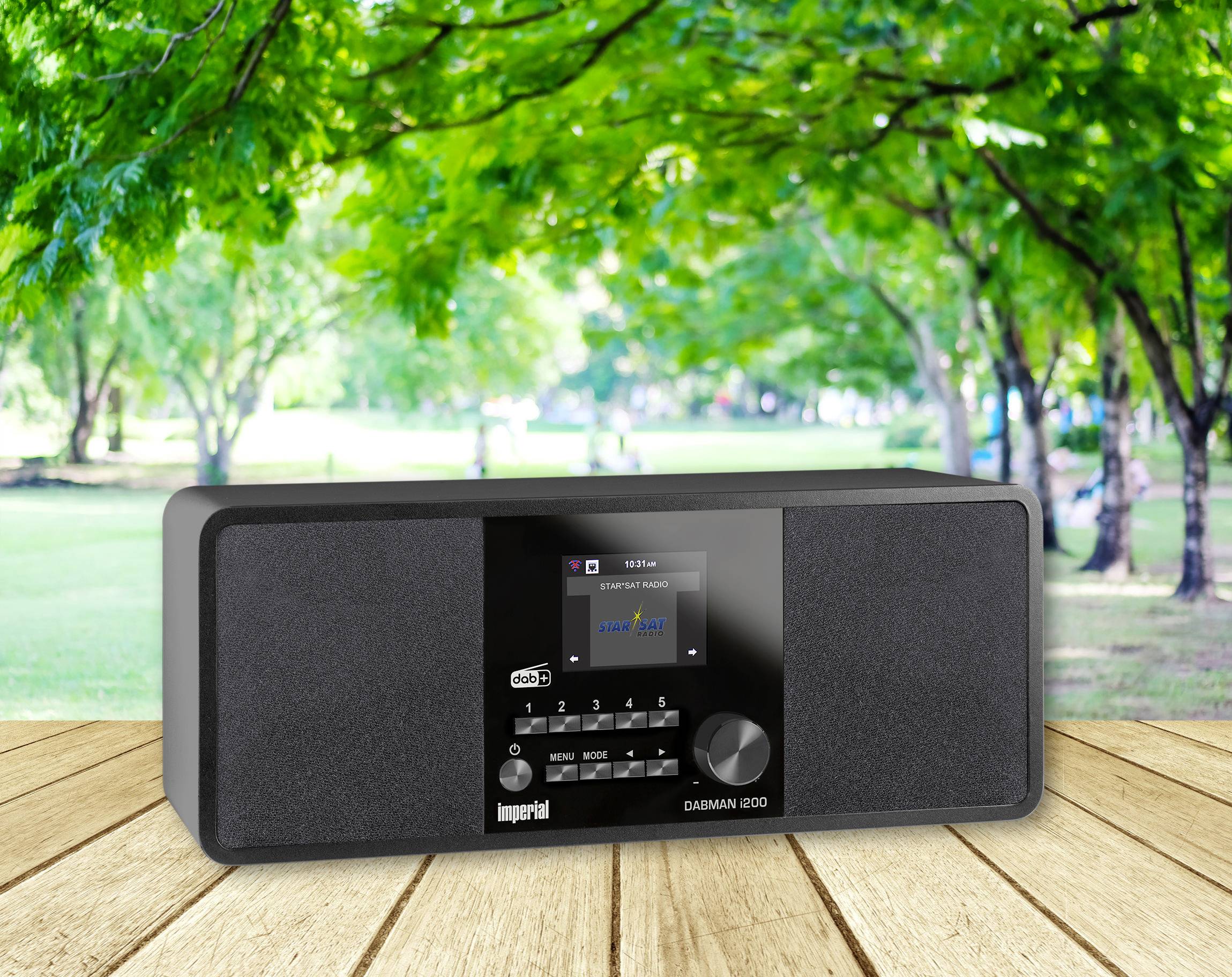 Ein Radio steht auf einem Holztisch vor einer grünen Parklandschaft. Es zeigt einen Musiksender auf dem Display an.