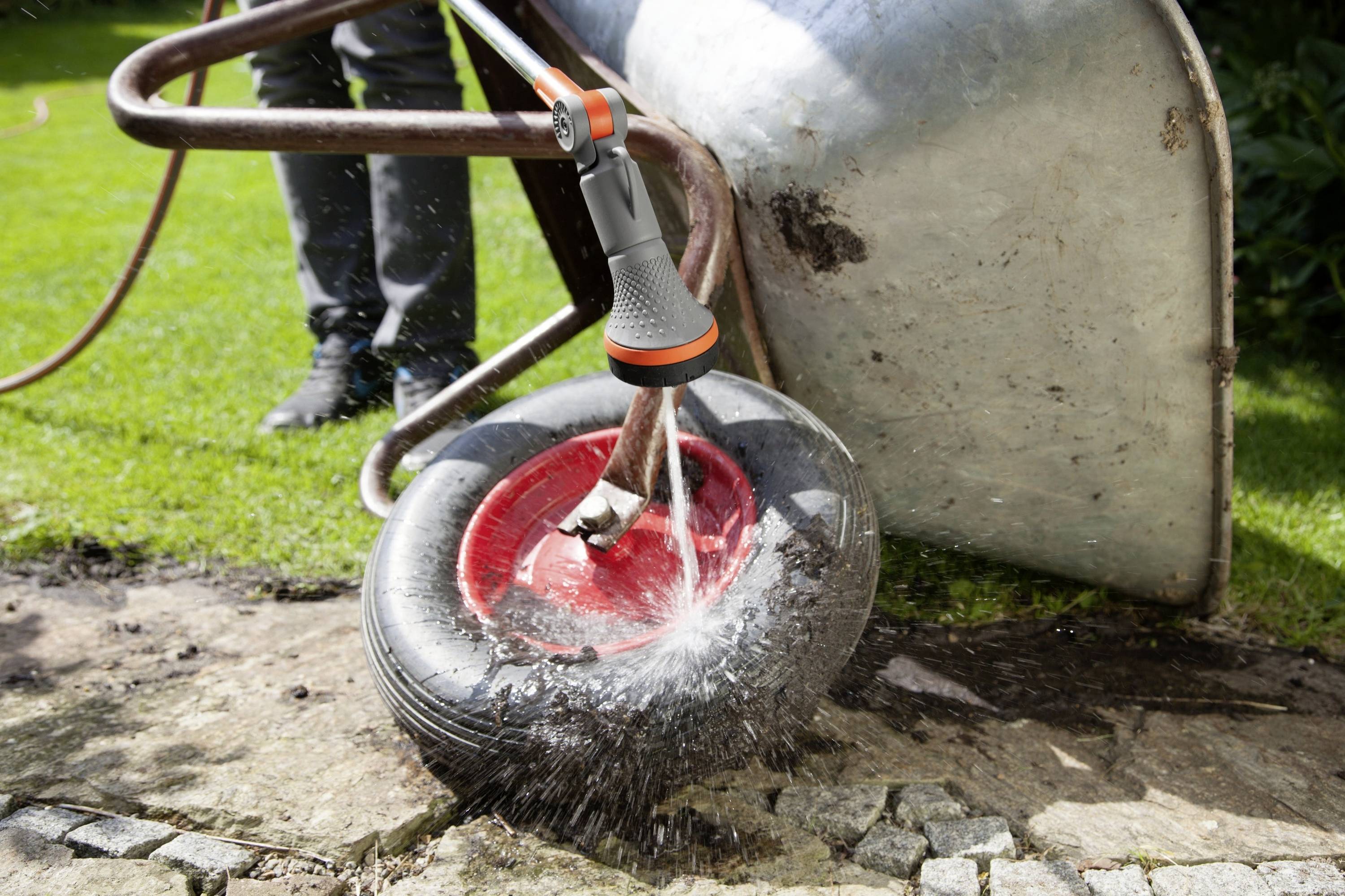 Eine Person spritzt mit einem Gartenschlauch den Reifen einer umgedrehten Schubkarre ab. Wasser trifft den roten Reifen auf Steinboden.