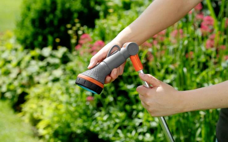 Eine Person justiert eine Gartenschlauchbrause vor einem Hintergrund mit einer Mischung aus grünen Pflanzen und blühenden Blumen.