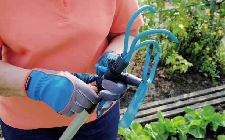 Eine Person mit Gartenhandschuhen befestigt ein Werkzeug an einem Griff und bereitet sich auf Gartenarbeit in der Nähe eines Gemüsegartens mit grünen Pflanzen vor.