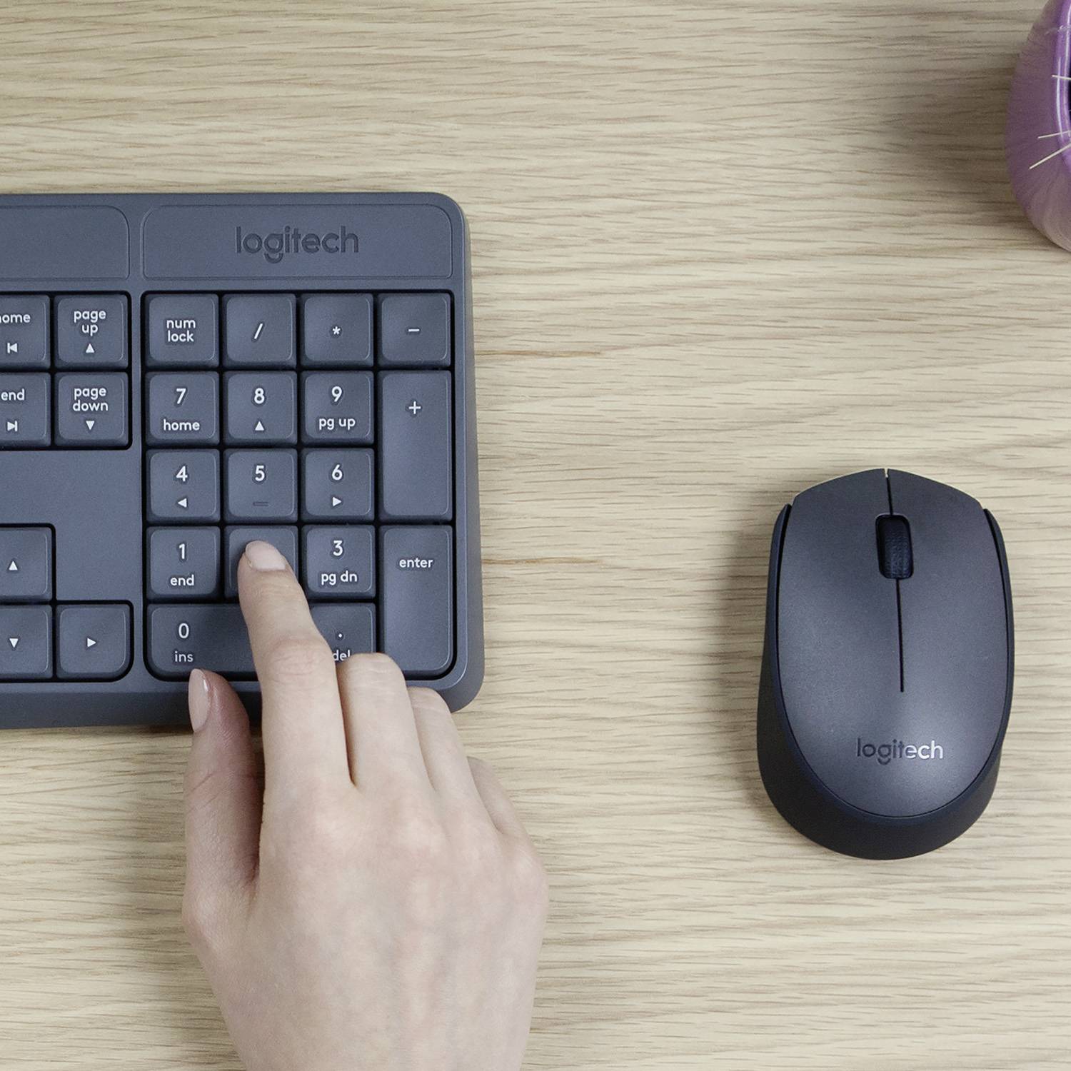 Eine Hand tippt auf eine Logitech-Zifferntastatur neben einer Logitech-Maus auf einem Holztisch.