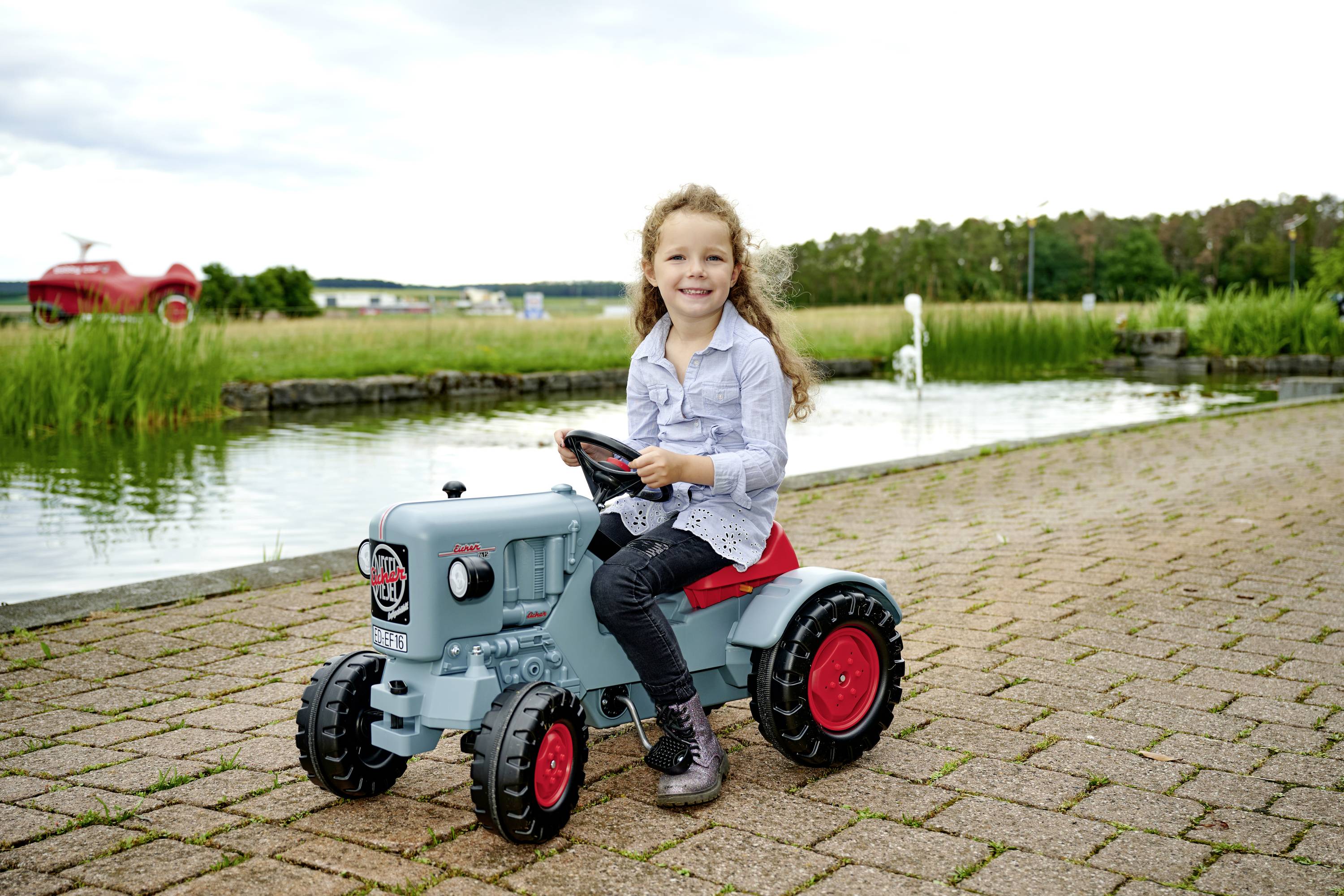 Ein lächelndes Kind sitzt auf einem Spielsachen-Oldtimer-Traktor auf einem gepflasterten Weg, mit einem Teich und Bäumen im Hintergrund.
