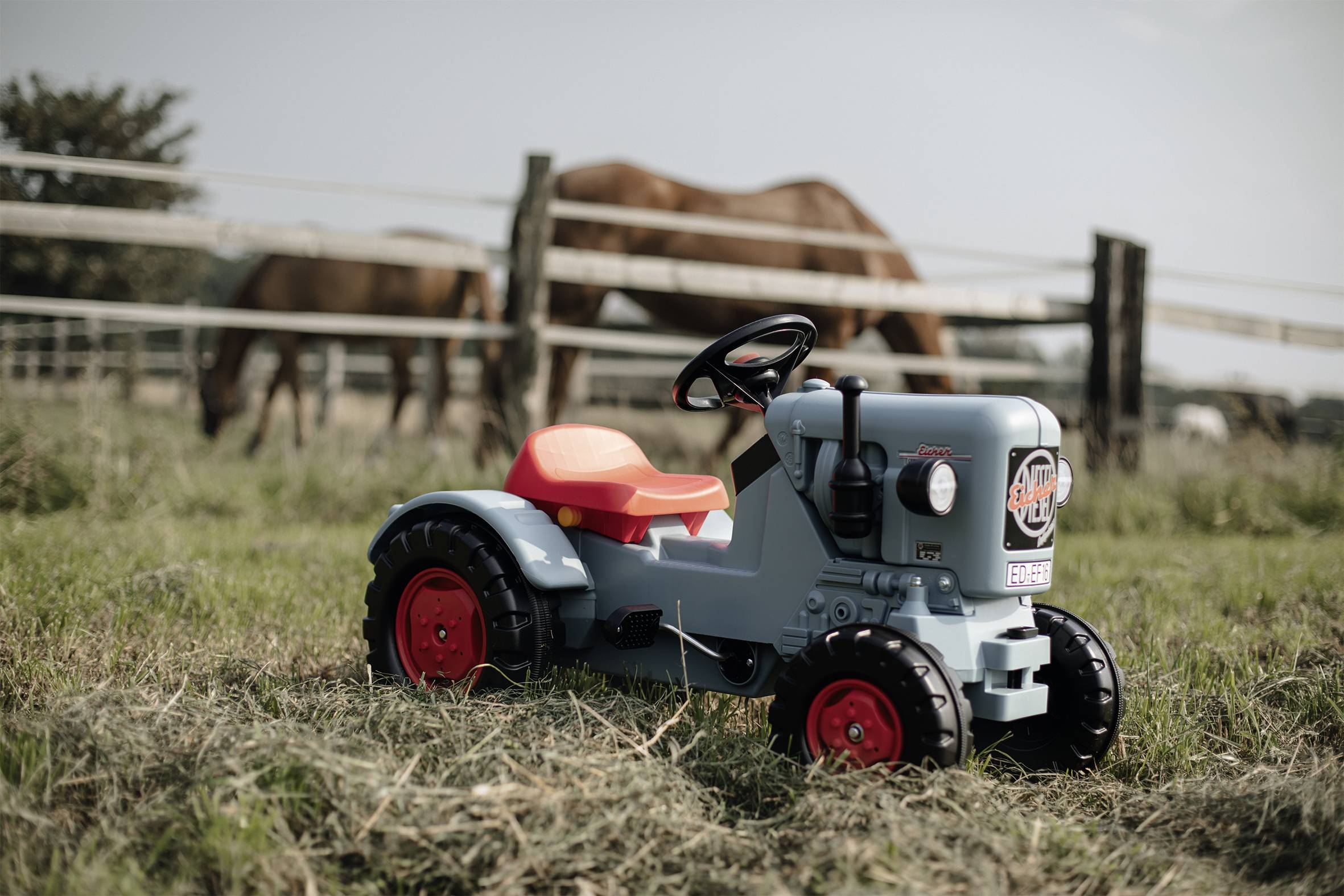 Ein Kinderspielzeugtraktor steht auf einer Wiese, im Hintergrund weiden Pferde hinter einem Zaun.