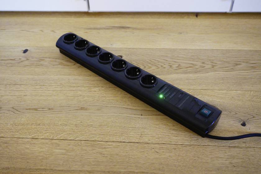 Eine schwarze Steckdosenleiste mit sieben Steckplätzen liegt auf einem Holzboden. Eine grüne Kontrollleuchte ist eingeschaltet.