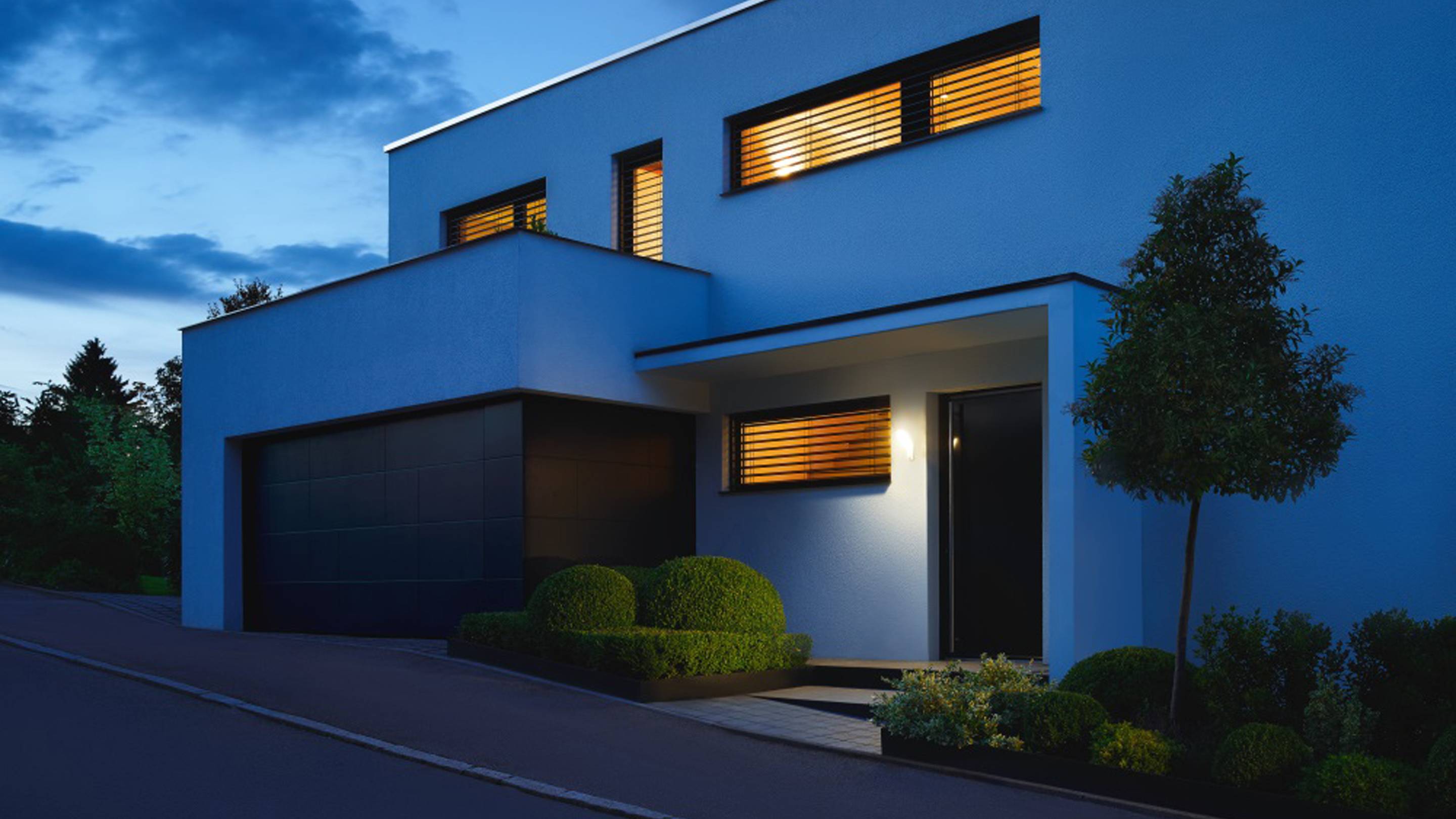 Modernes zweistöckiges Haus bei Dämmerung, beleuchtete Fenster, gepflegter Garten, Garage links.