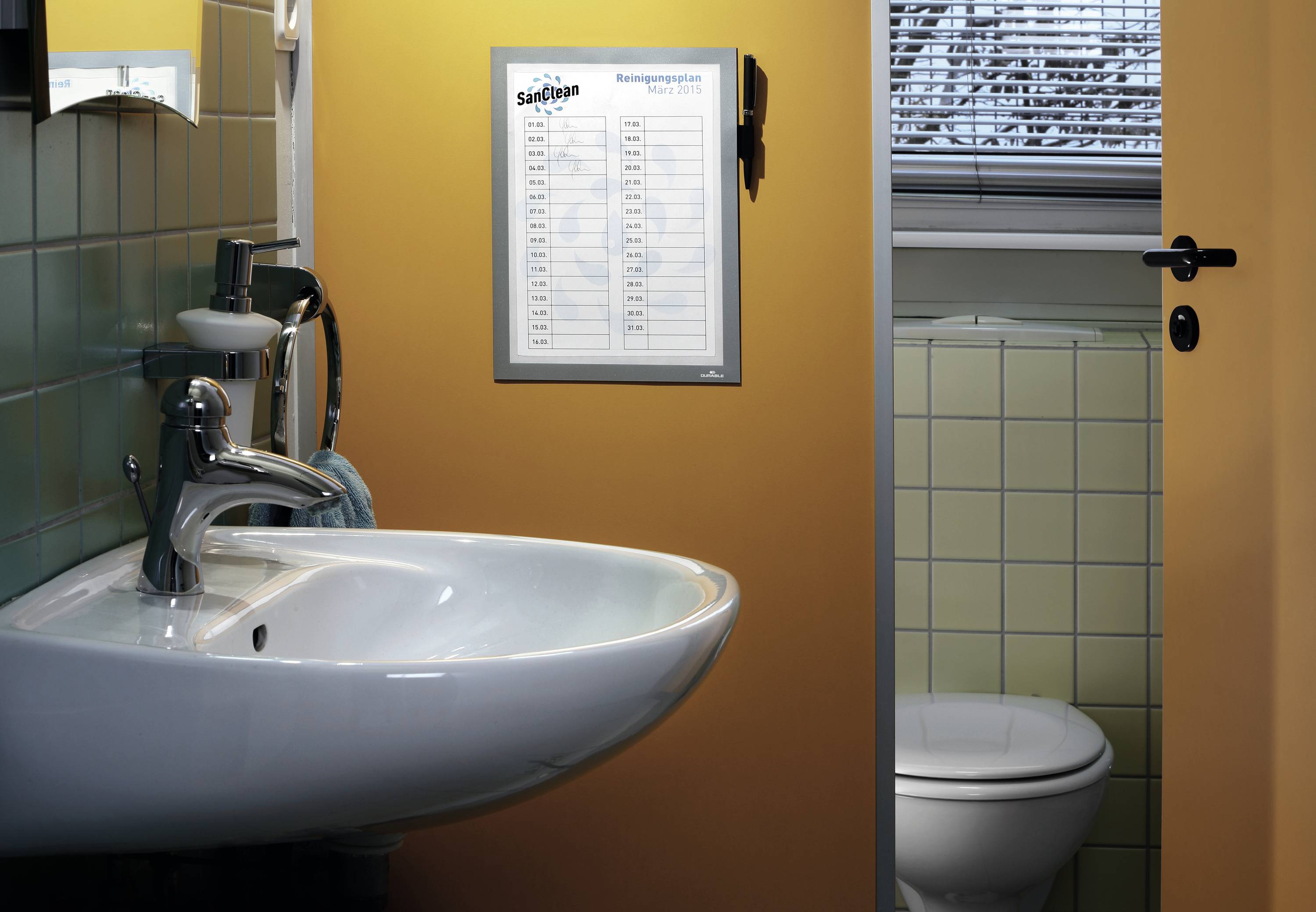 Waschbecken und geöffnete Badezimmertür mit Blick auf Toilette; Reinigungsplan an der Wand über dem Waschbecken.