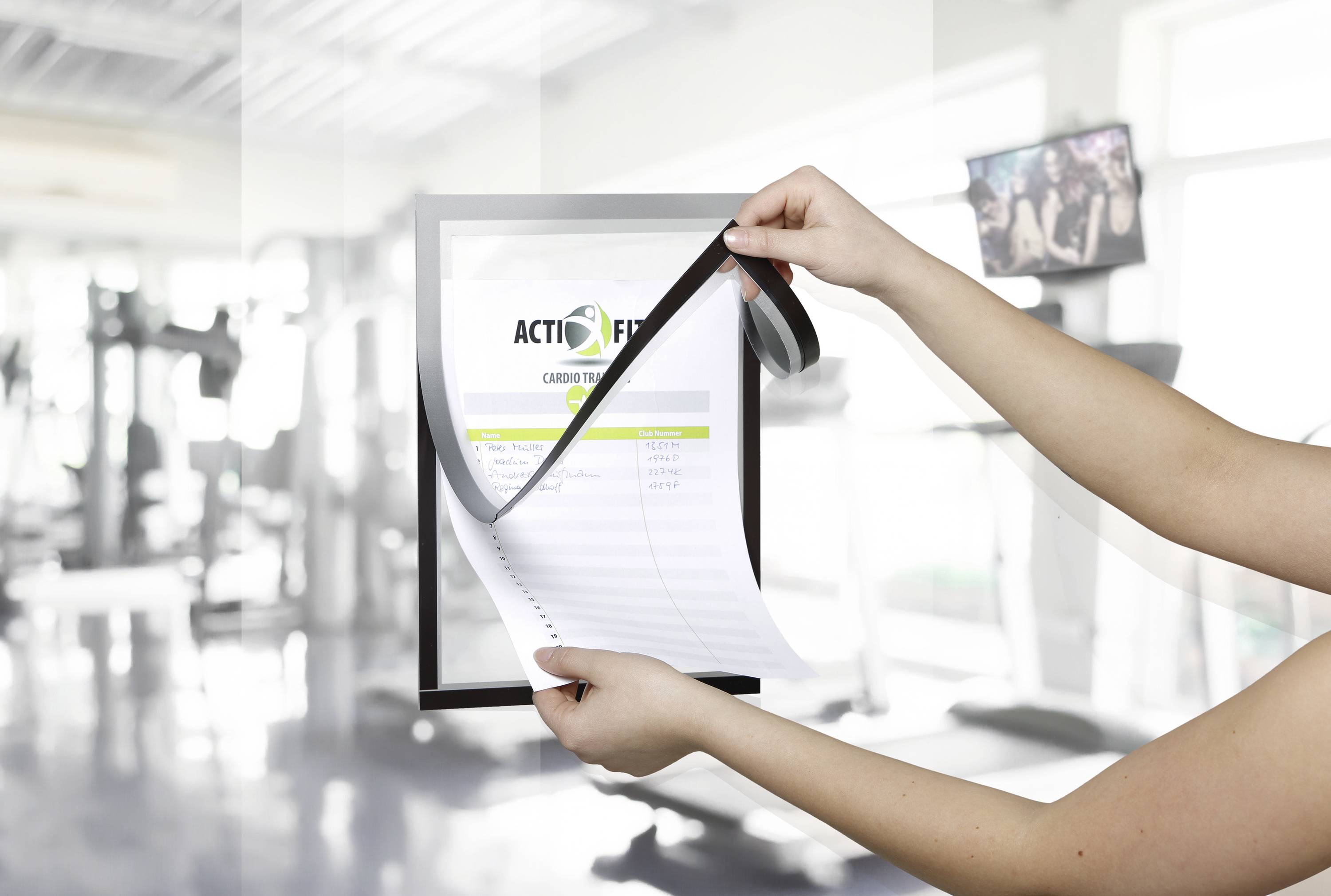 Person hält ein Fitness-Plan-Heft in einem Fitnessstudio, Hintergrund zeigt verschwommene Trainingsgeräte.