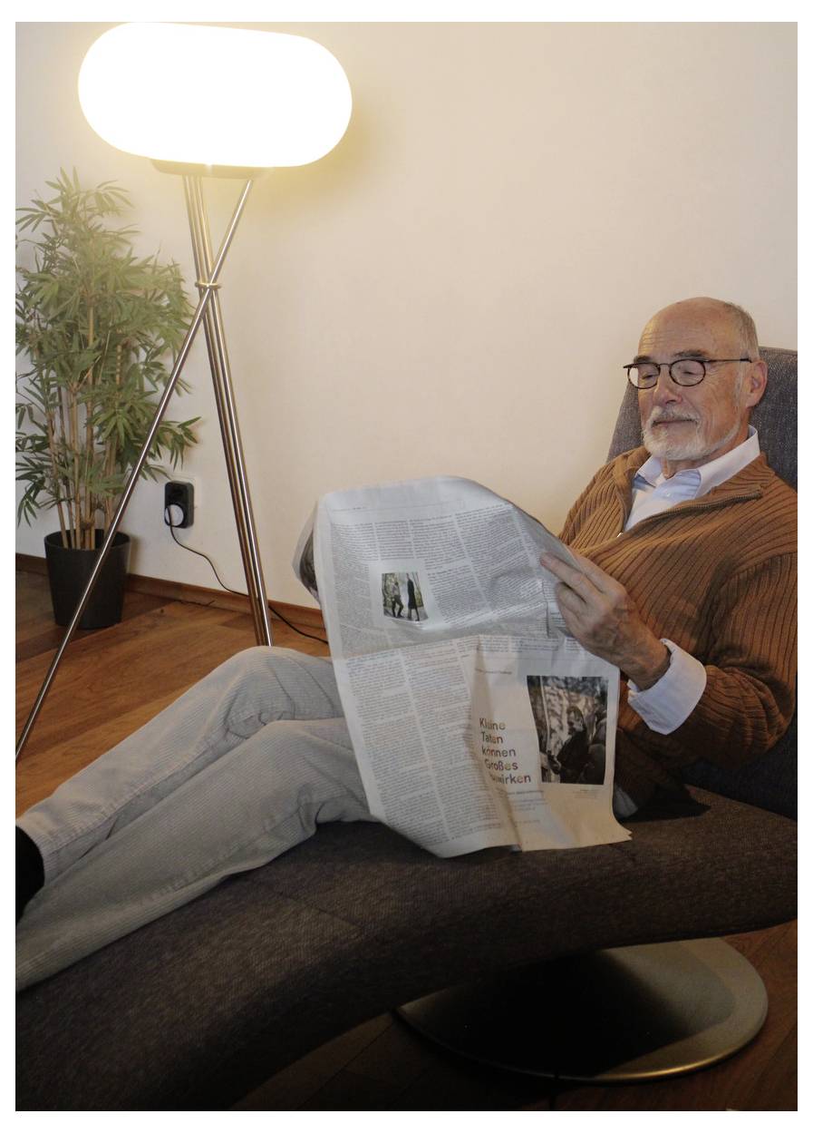 Ein älterer Mann mit Brille entspannt sich in einem Sessel und liest eine Zeitung neben einer hellen Stehleuchte und einer Topfpflanze.