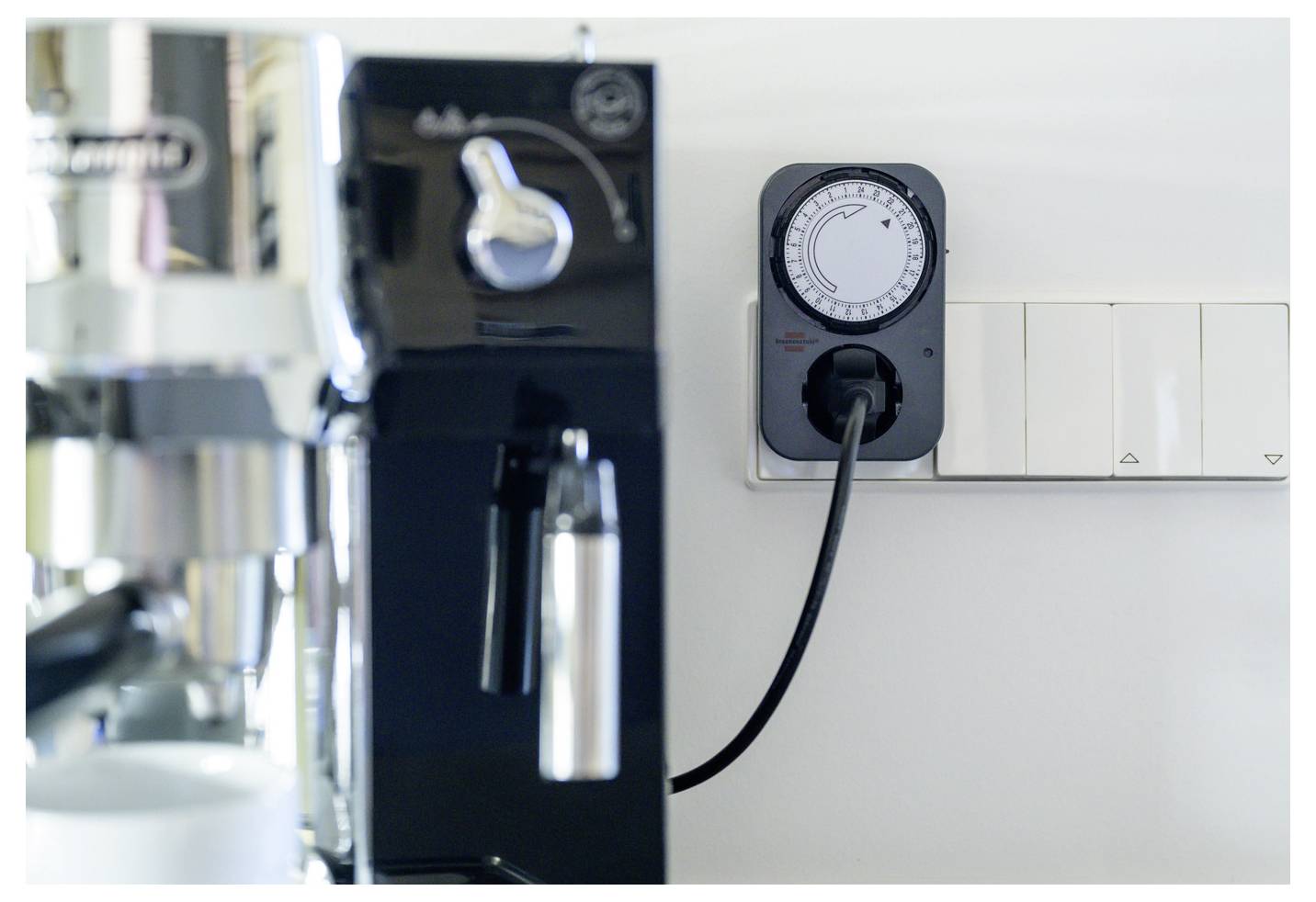 Wandmontierter Timer-Schalter mit angeschlossenem Kabel neben einer Kaffeemaschine in einer Küche oder einem Pausenraum.