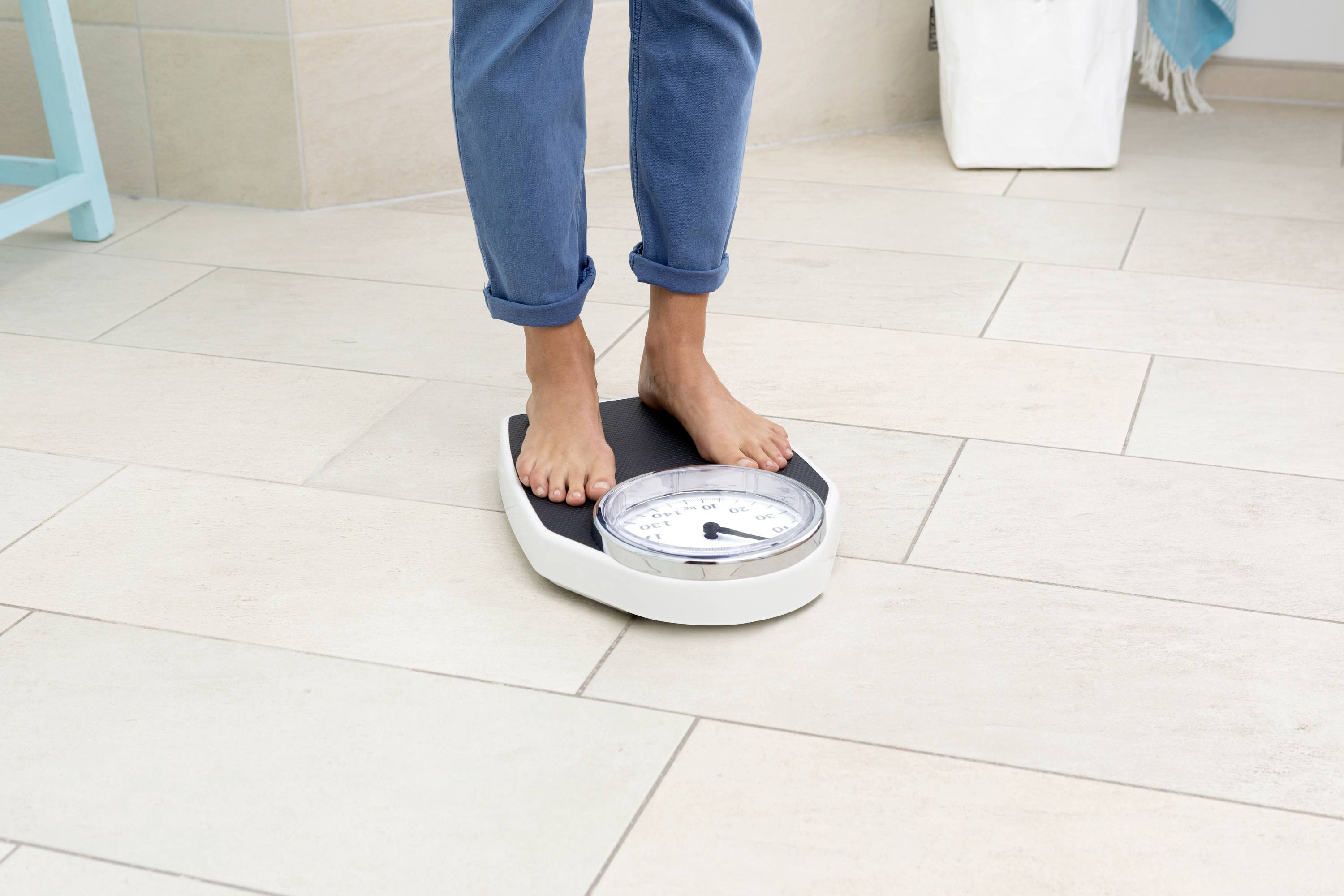 Person steht auf einer analogen Waage auf hellen Fliesen, trägt blaue Jeans.