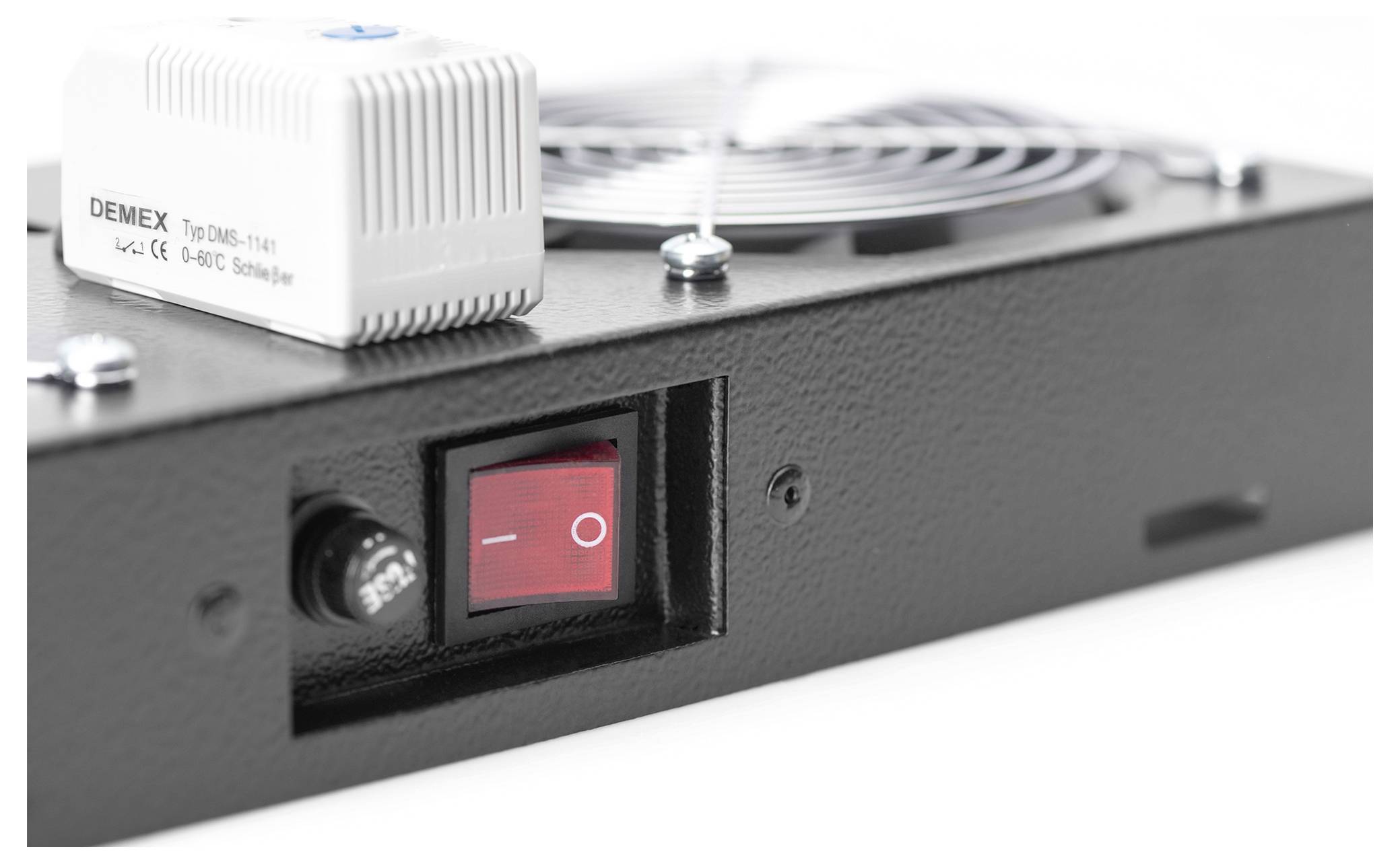 Eine Nahaufnahme eines schwarzen elektronischen Geräts mit einem roten Ein-/Ausschalter und einer weißen DEMEX-Komponente mit einem Lüftungsgitter im Hintergrund.