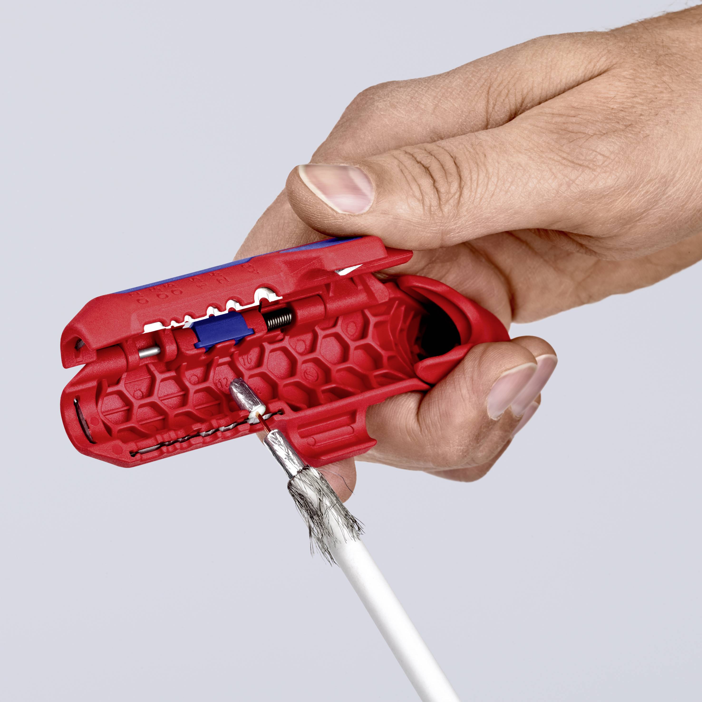 Eine Hand hält ein rotes Werkzeug, das gerade ein Kabel abisoliert. Das Werkzeug hat eine wabenförmige Struktur im Griffbereich.