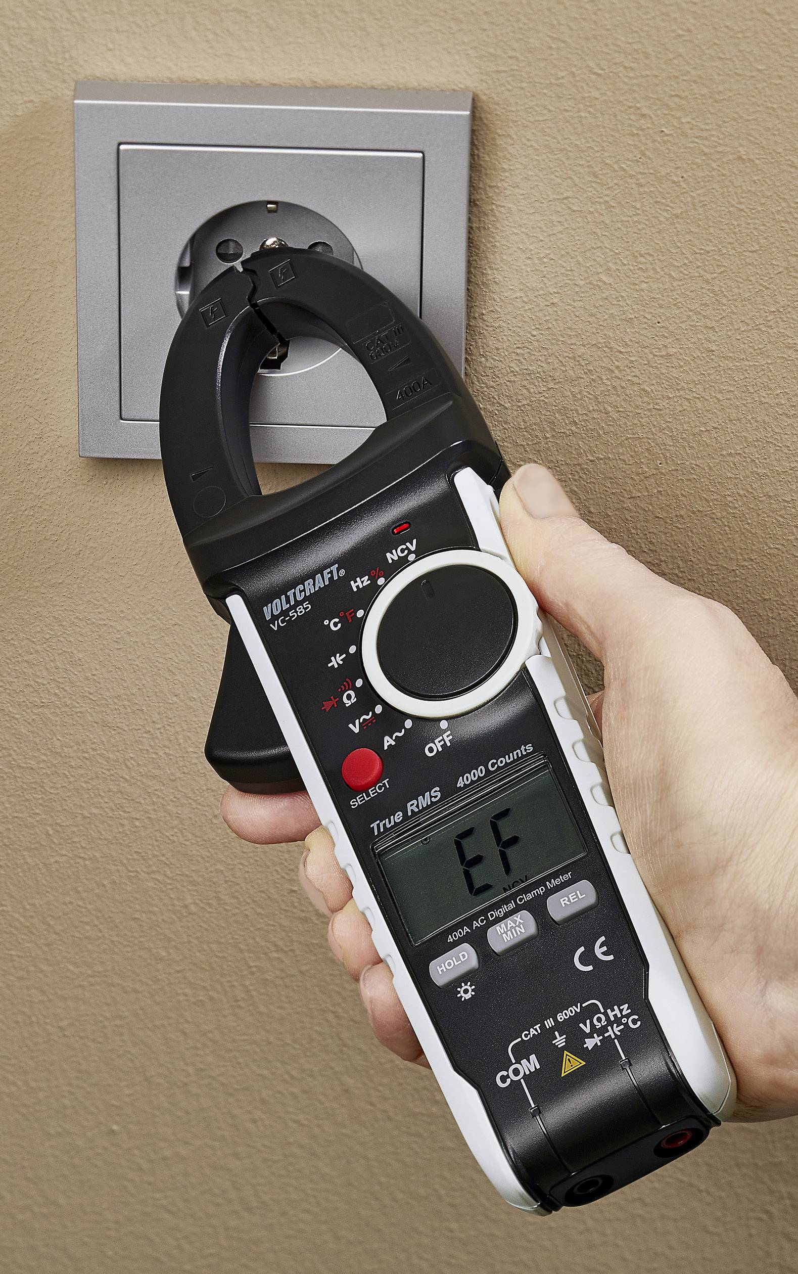 Eine Hand hält ein Zangenamperemeter, das an eine Steckdose angeschlossen ist, um elektrische Messungen durchzuführen.