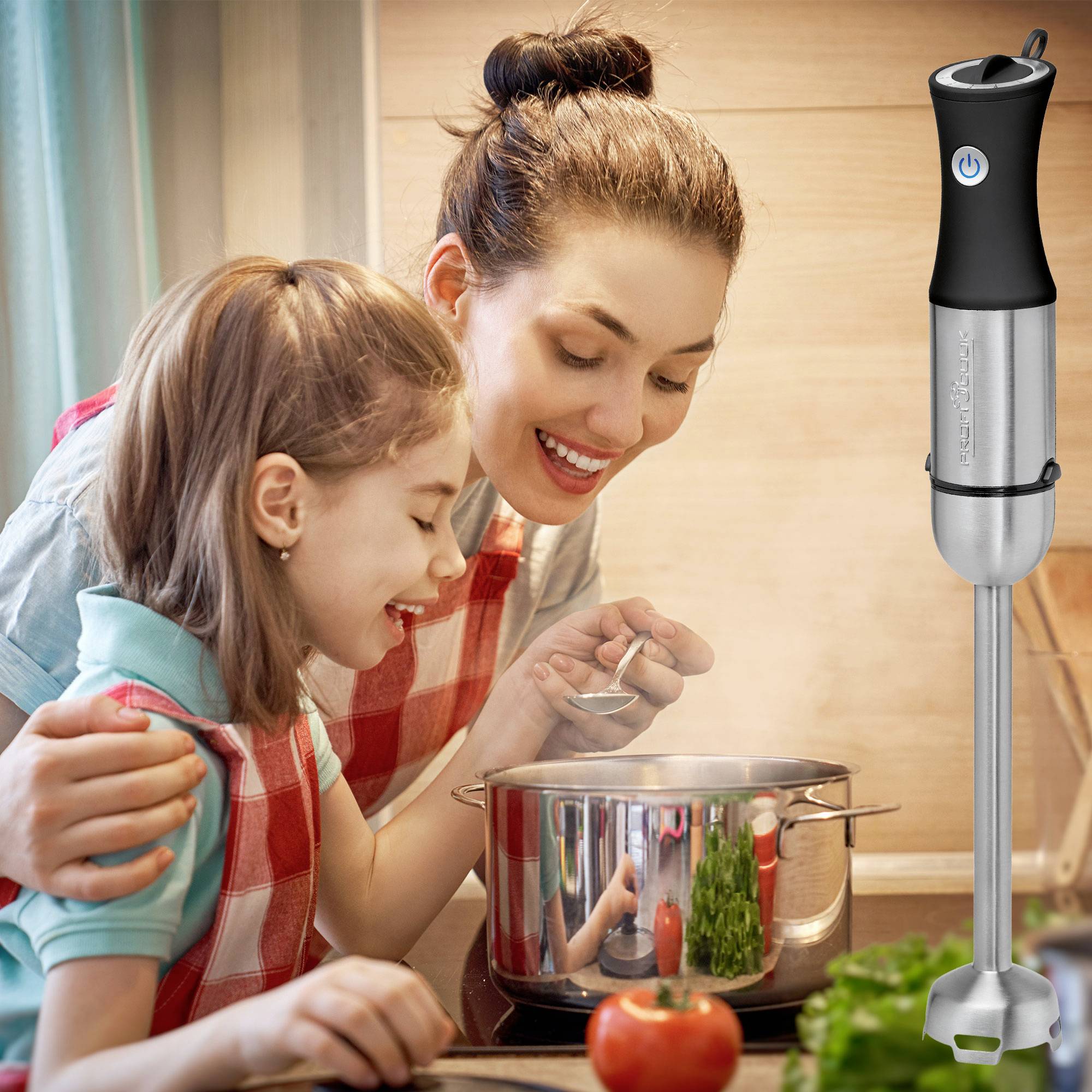 Mutter und Tochter kochen zusammen in der Küche und sehen in einen Topf. Ein Stabmixer steht links von ihnen auf der Arbeitsfläche.