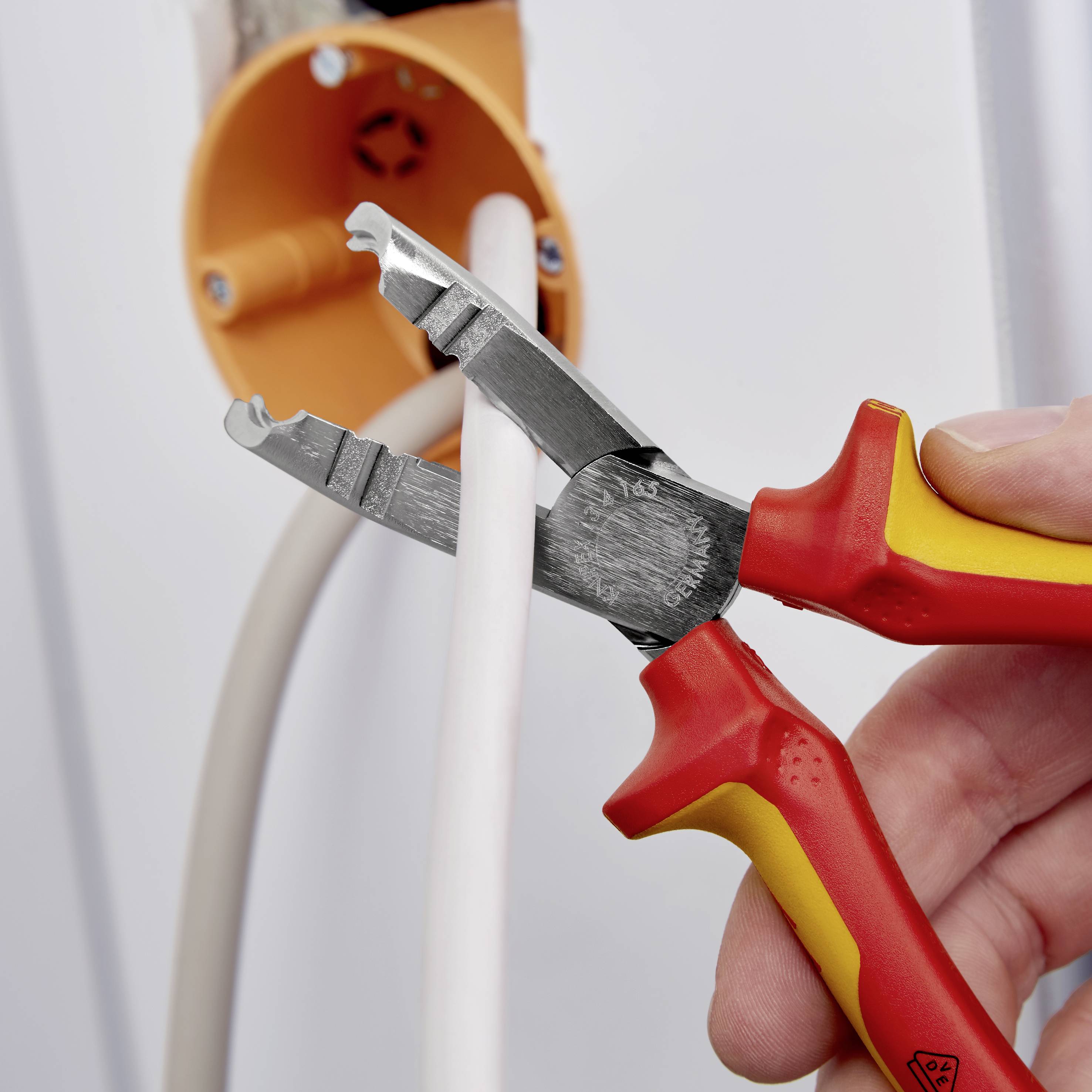 Eine Hand hält eine rote Zange und schneidet ein elektrisches Kabel in einer orangefarbenen Steckdose.
