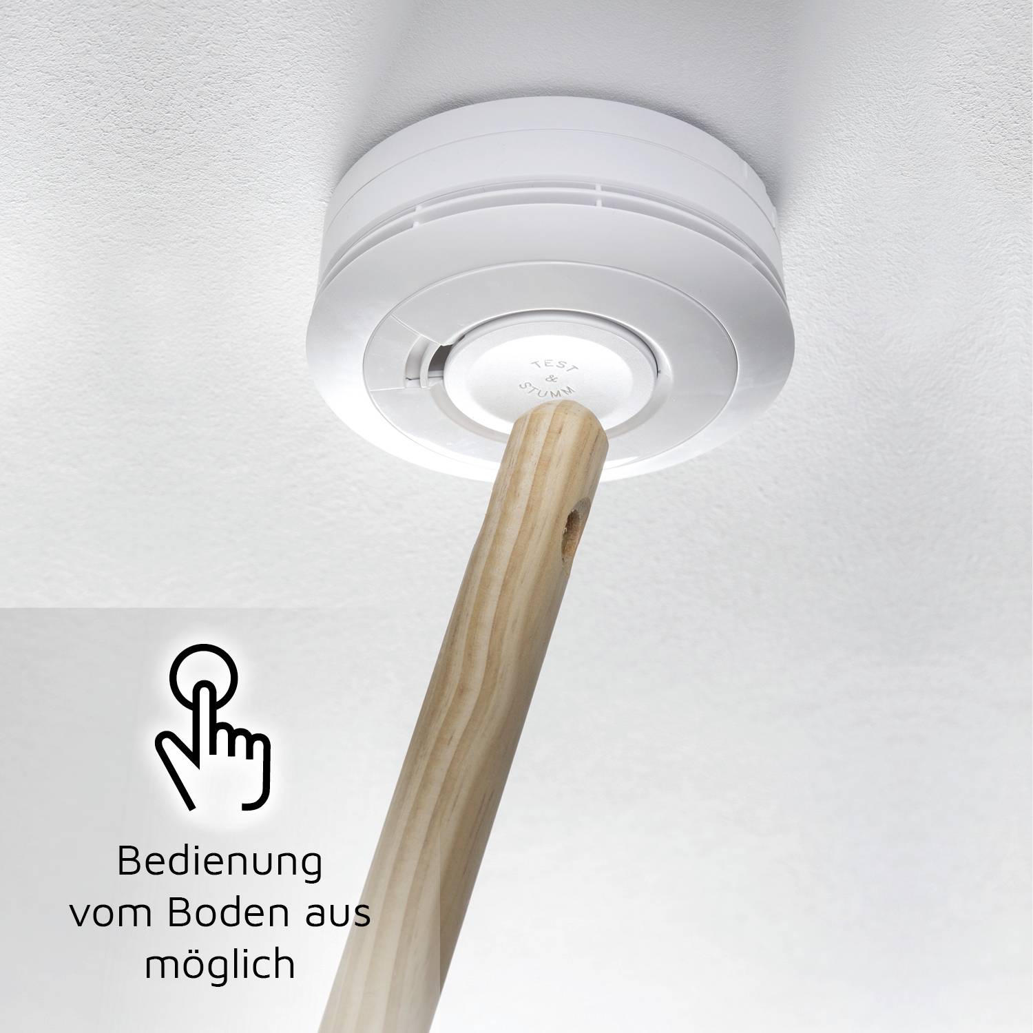 Ein Rauchmelder an der Decke wird mit einem Holzstab bedient. Ein Symbol daneben zeigt einen Finger und der Text lautet 'Bedienung vom Boden aus möglich'.