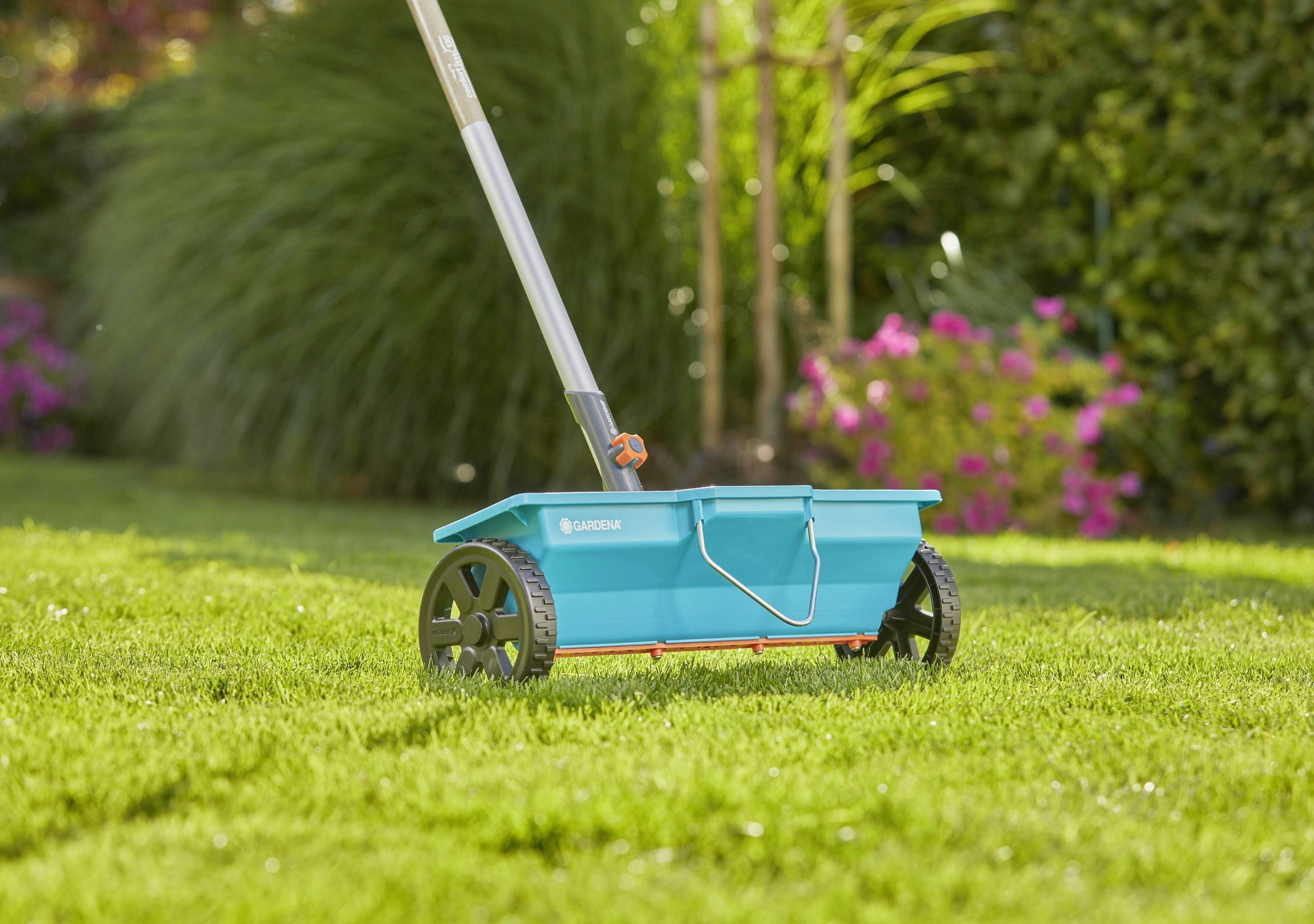 Ein blauer Streuwagen auf einer grünen Rasenfläche, umgeben von Blumen und Büschen im Hintergrund.
