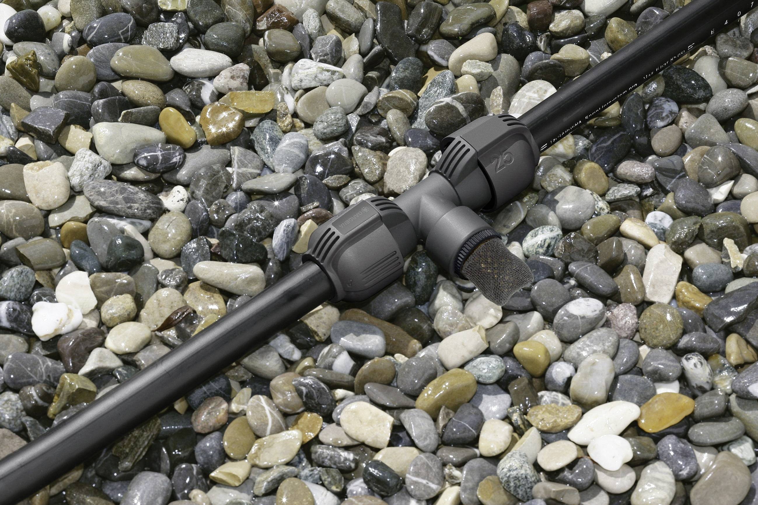 Ein Bewässerungsrohr mit T-Stück auf einem Untergrund aus bunten Kieselsteinen, bereit für Gartenbewässerungssysteme.