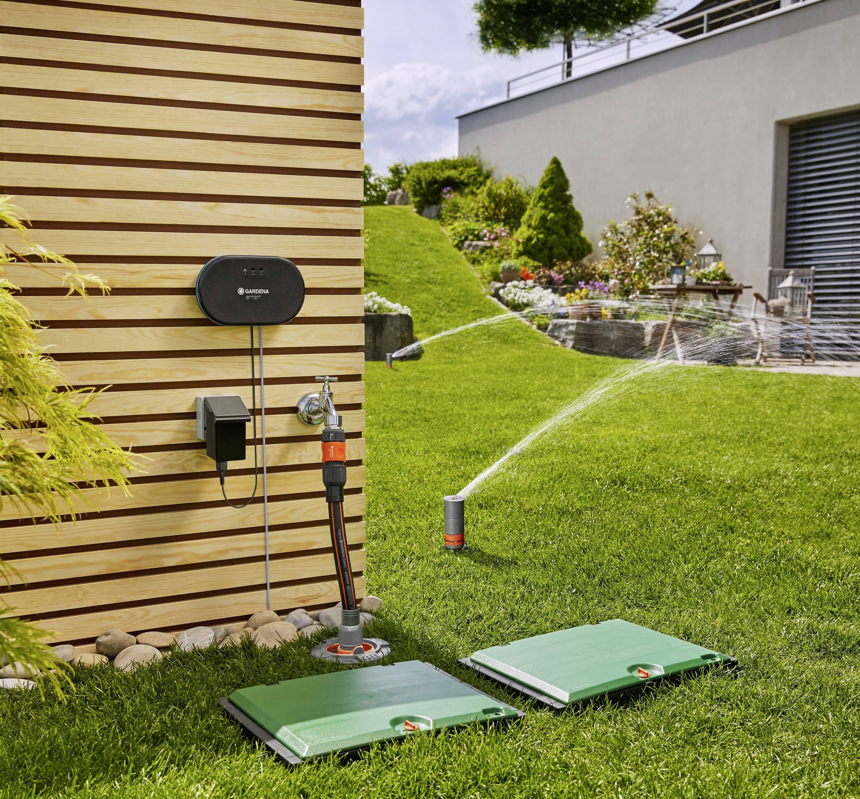 Automatisiertes Bewässerungssystem im Garten mit Sprinklern und Steuerungseinheit an holzverkleideter Wand, Rasen und Blumenbeet.