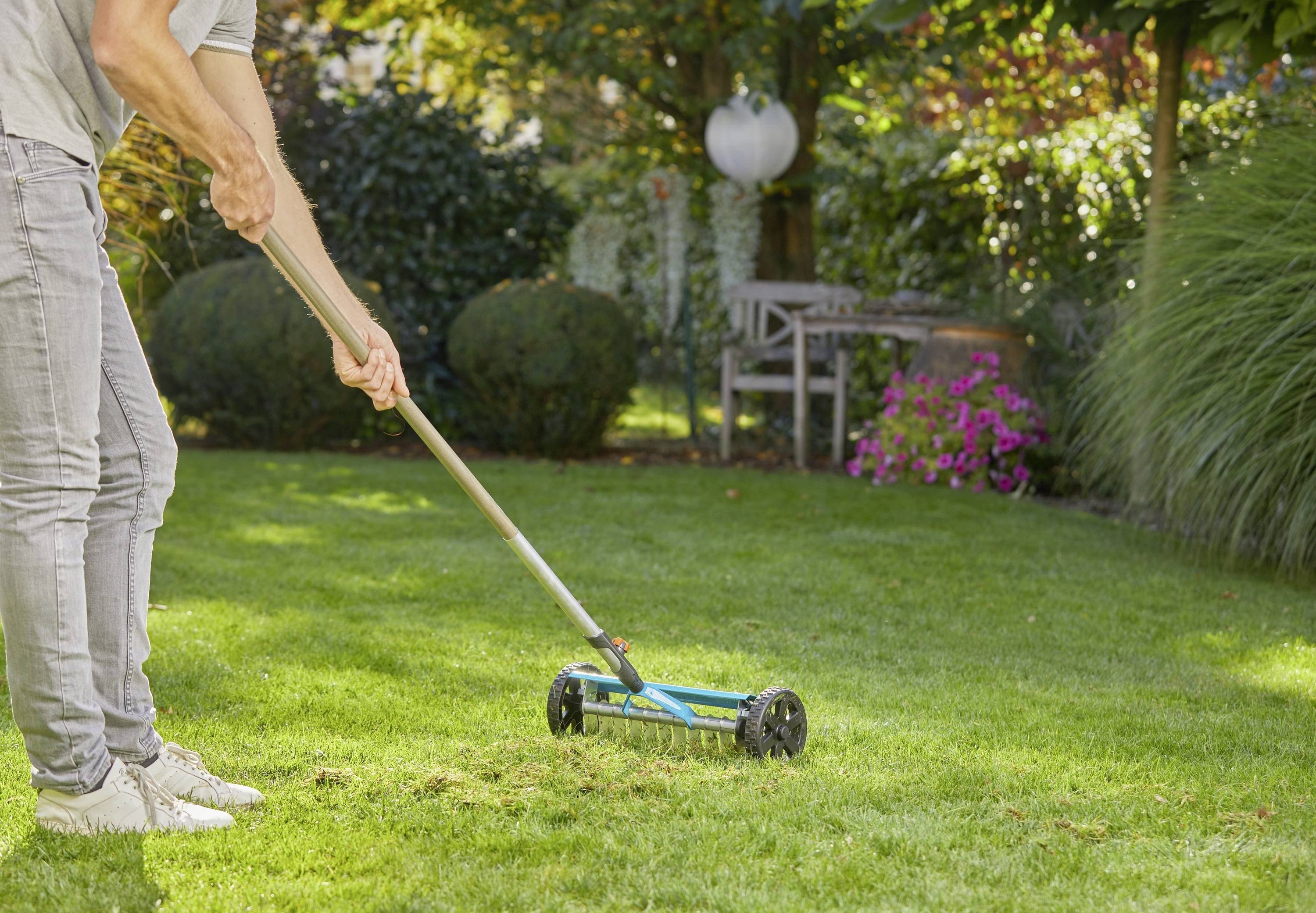 Eine Person mäht mit einem Handrasenmäher das Gras in einem Garten. Im Hintergrund sind Büsche und Blumen zu sehen.