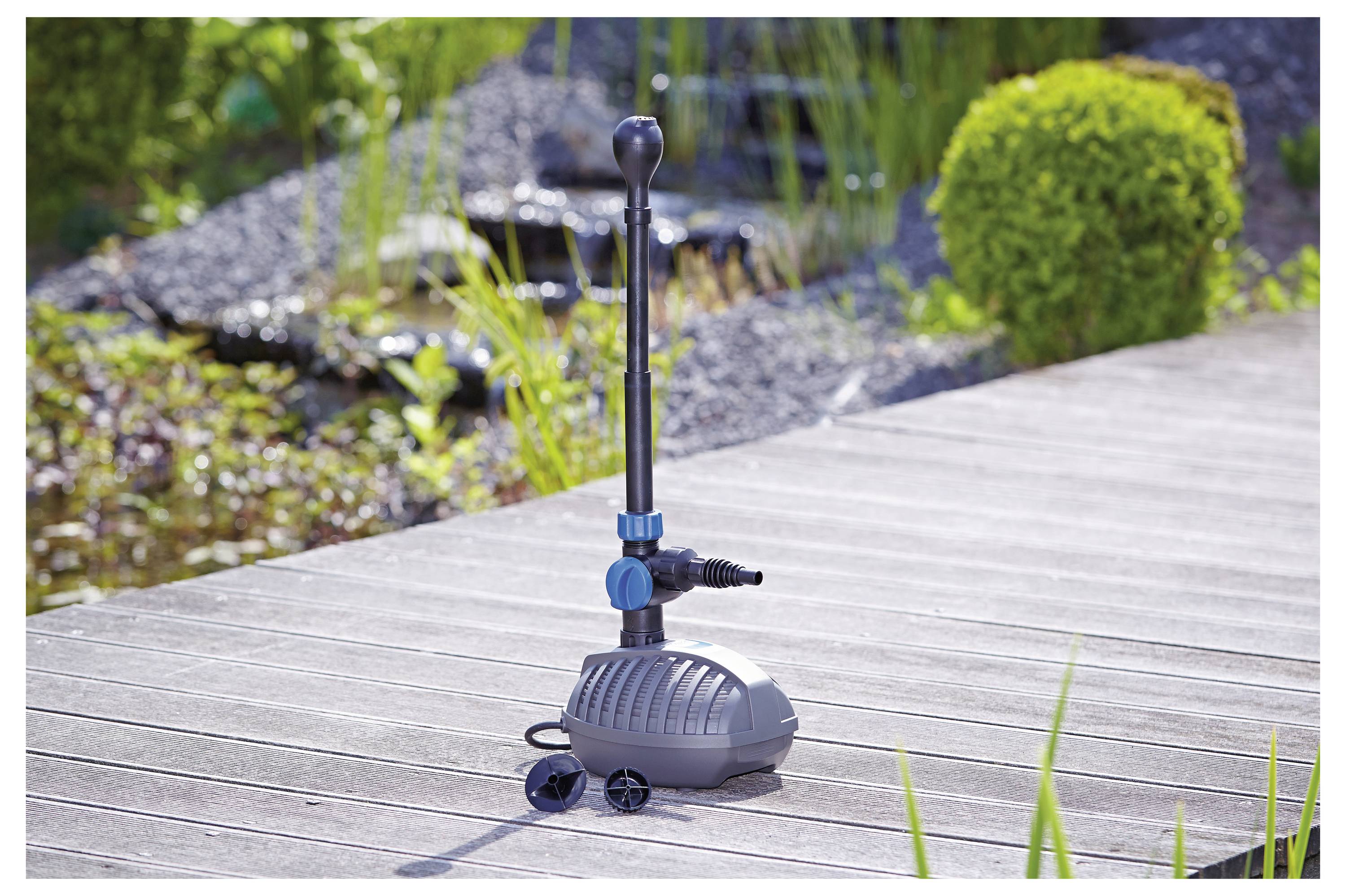 Eine Gartenbrunnen-Pumpe steht auf einer Holzterrasse in der Nähe eines Teiches, umgeben von Grünpflanzen und Steinen. Die Pumpe verfügt über einstellbare Ventile und Anschlüsse.
