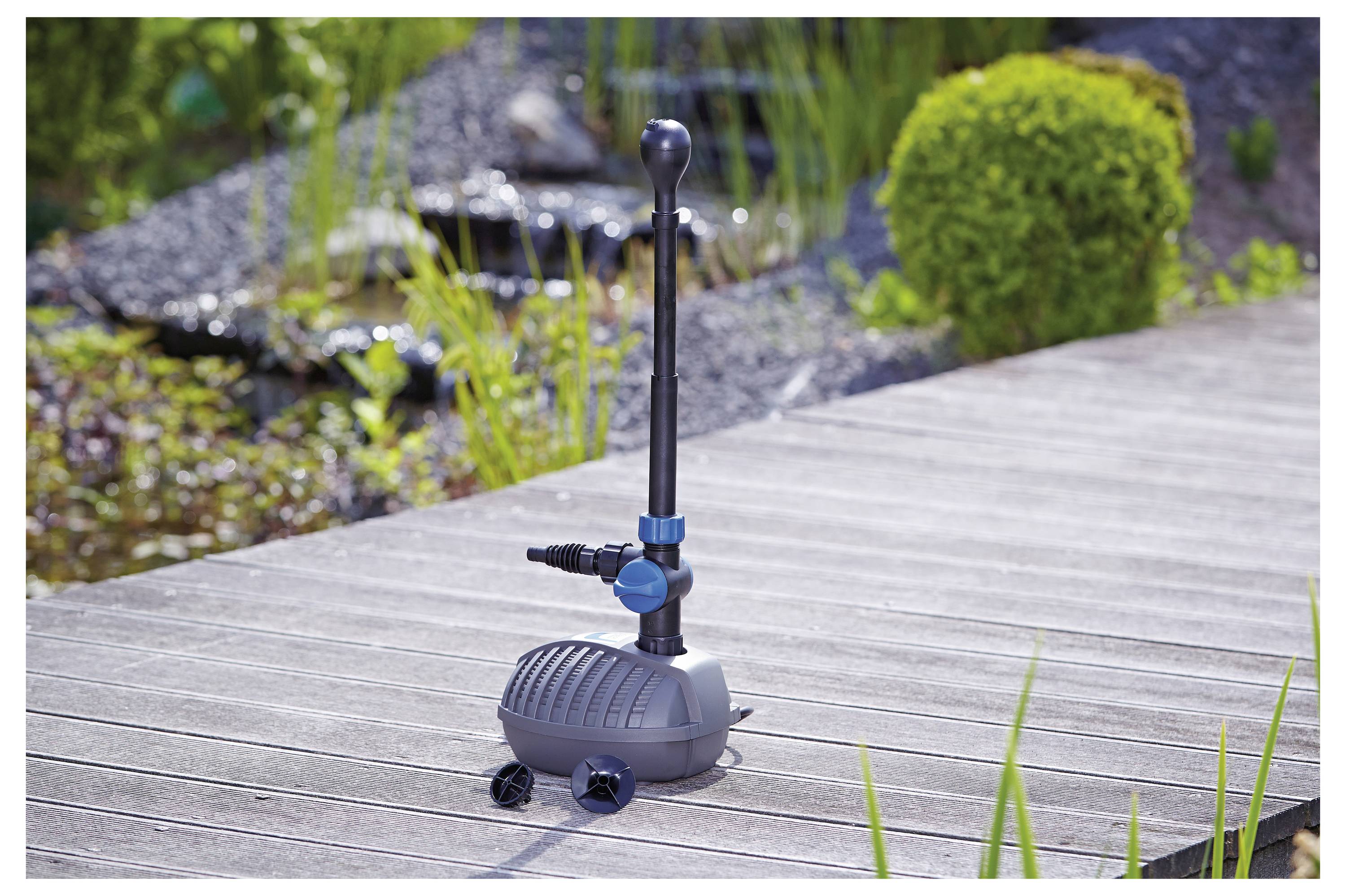 Ein kleiner Gartenbrunnen-Pumpe mit vertikalem Auslauf und blauer Ventilsteuerung steht auf einer Holzterrasse, umgeben von Grünpflanzen und Steinen.