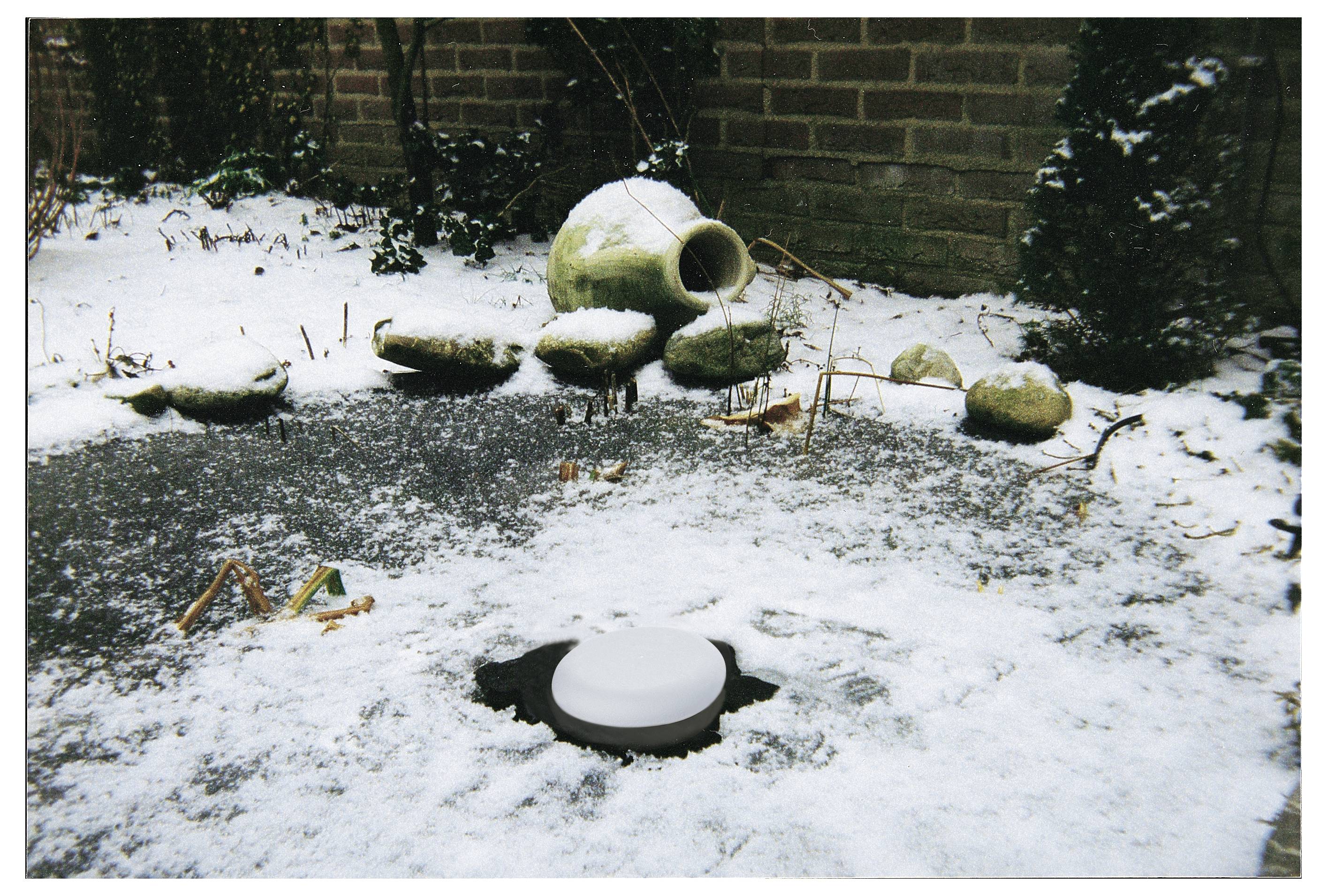 Ein schneebedeckter Garten mit verstreuten Steinen und einem runden, offenmündigen Tontopf im Hintergrund.