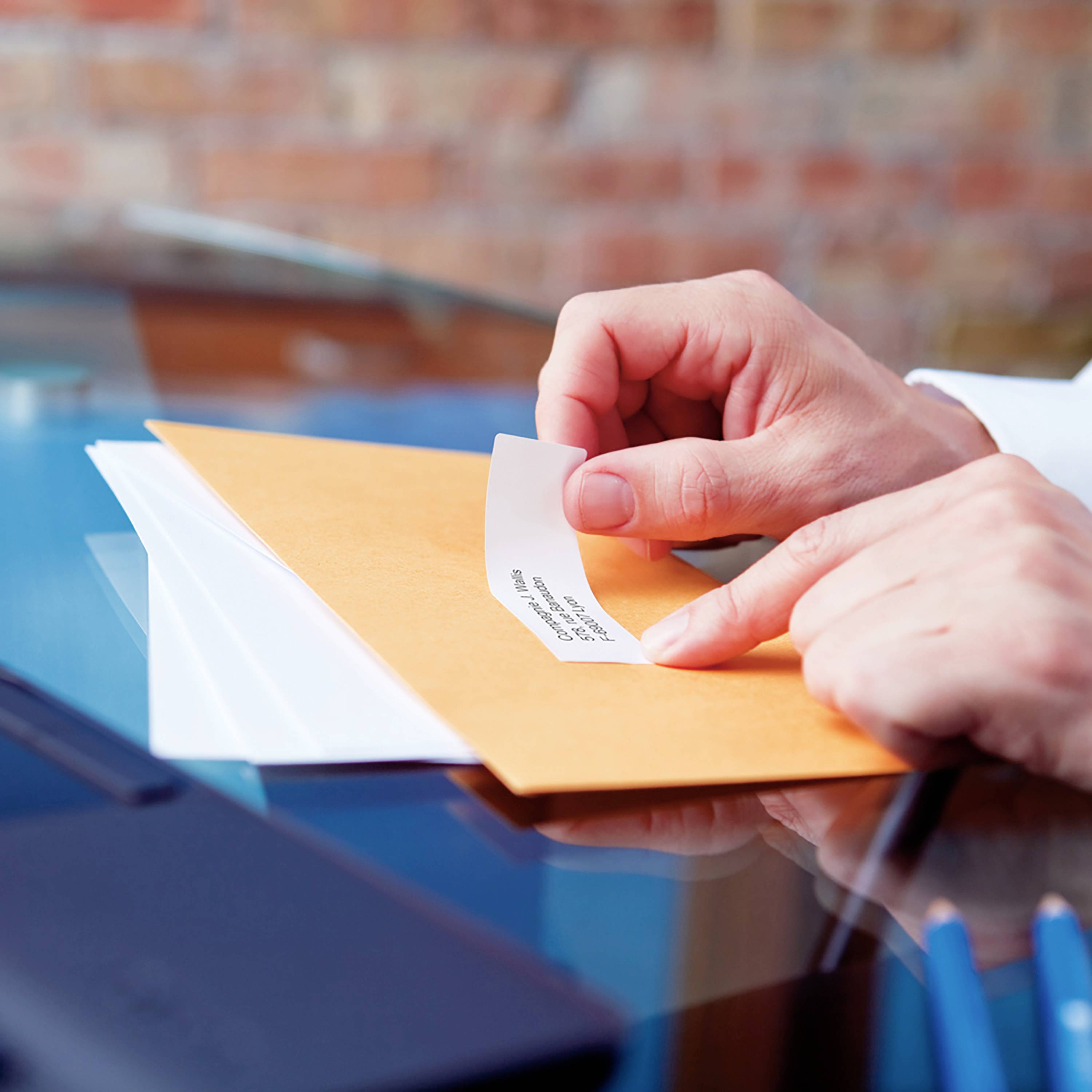 Eine Hand hält einen gelben Umschlag mit einem Etikett, auf dem Text steht. Im Hintergrund unscharf. Focus liegt auf dem Umschlag.